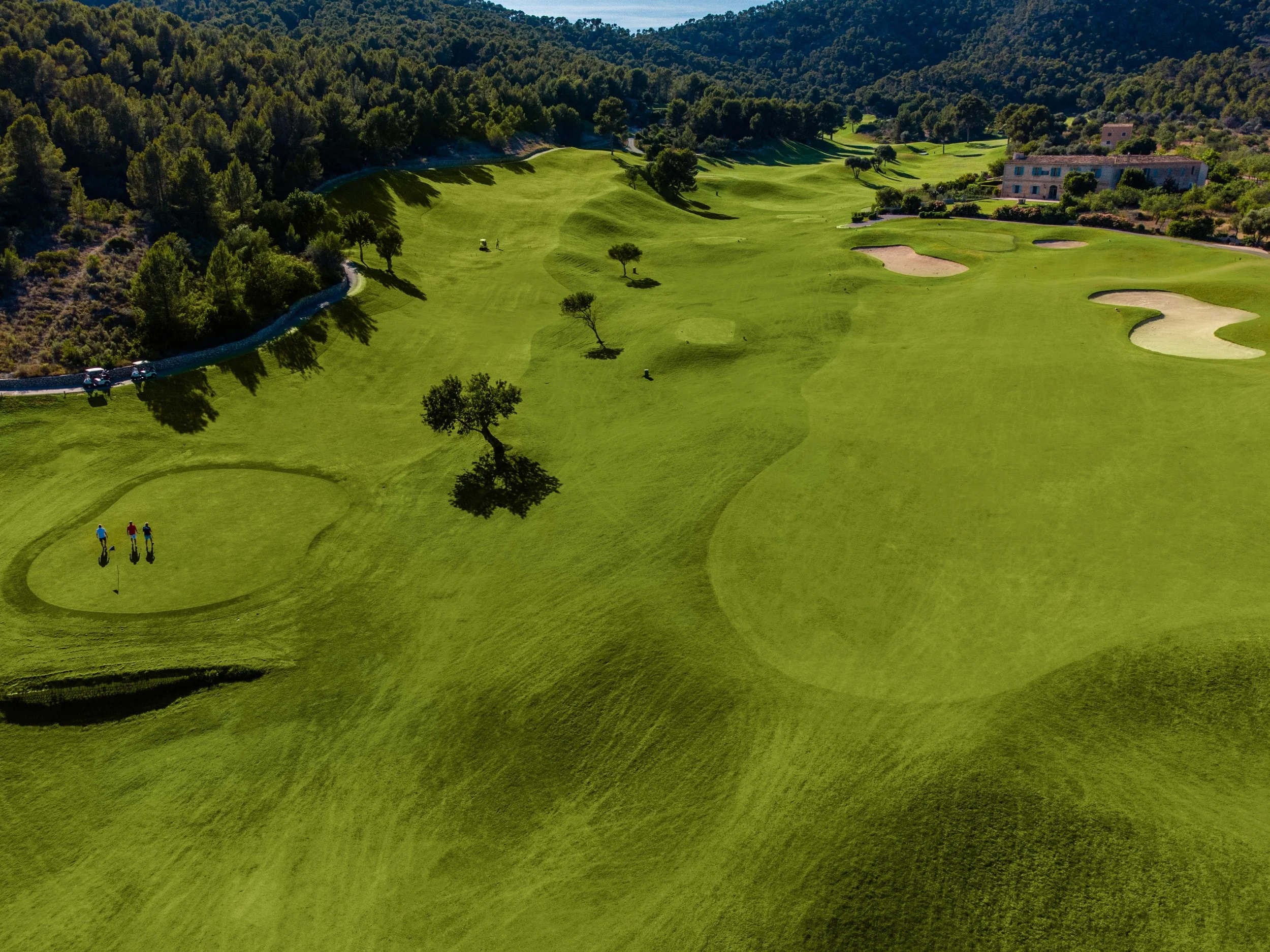 Vista aérea de un campo de golf con greens, árboles, y obstáculos de arena y agua en un paisaje verde y montañoso.