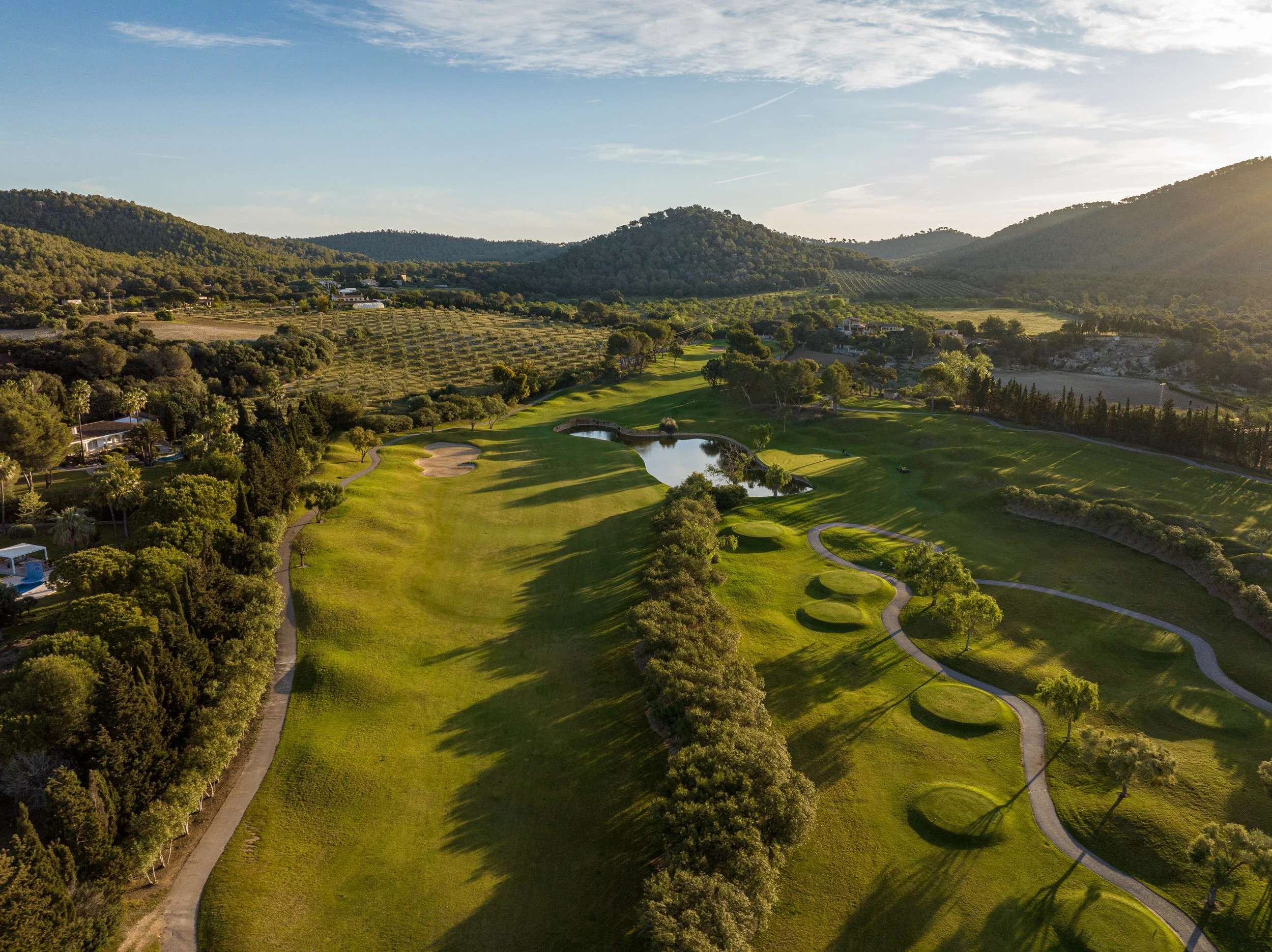 Vista aérea de un campo de golf rodeado de árboles, colinas y montañas, con un lago en el centro, durante el atardecer