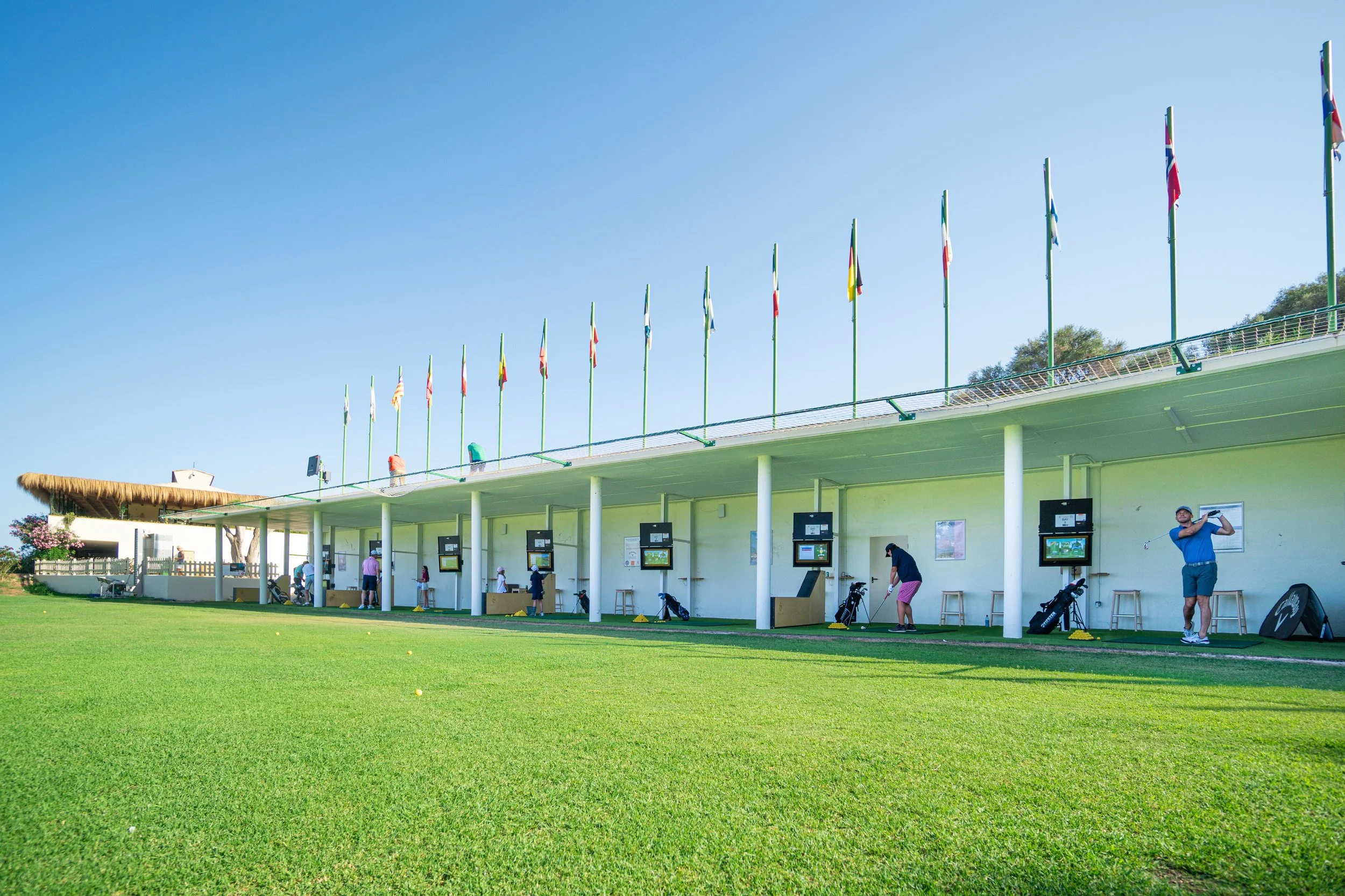 Nueve personas practicando golf en un campo al aire libre con palcos y banderas de diferentes países.