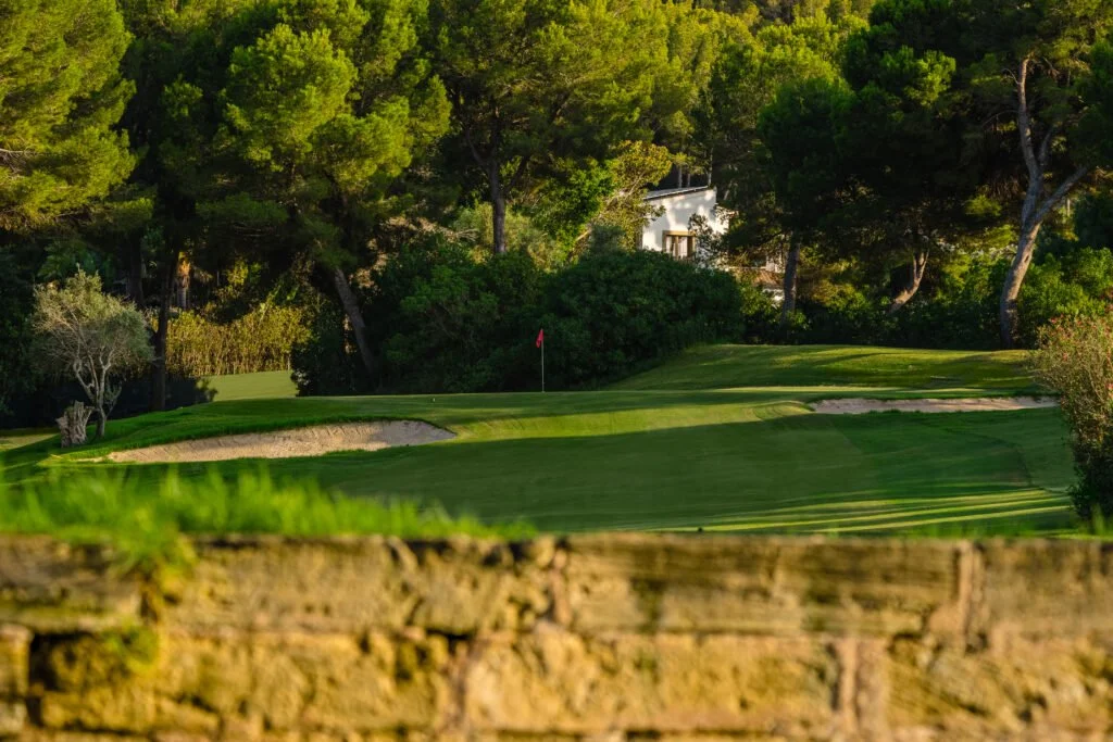 Vista de un campo de golf con verde césped, obstáculos de arena, árboles y una casa blanca en el fondo.