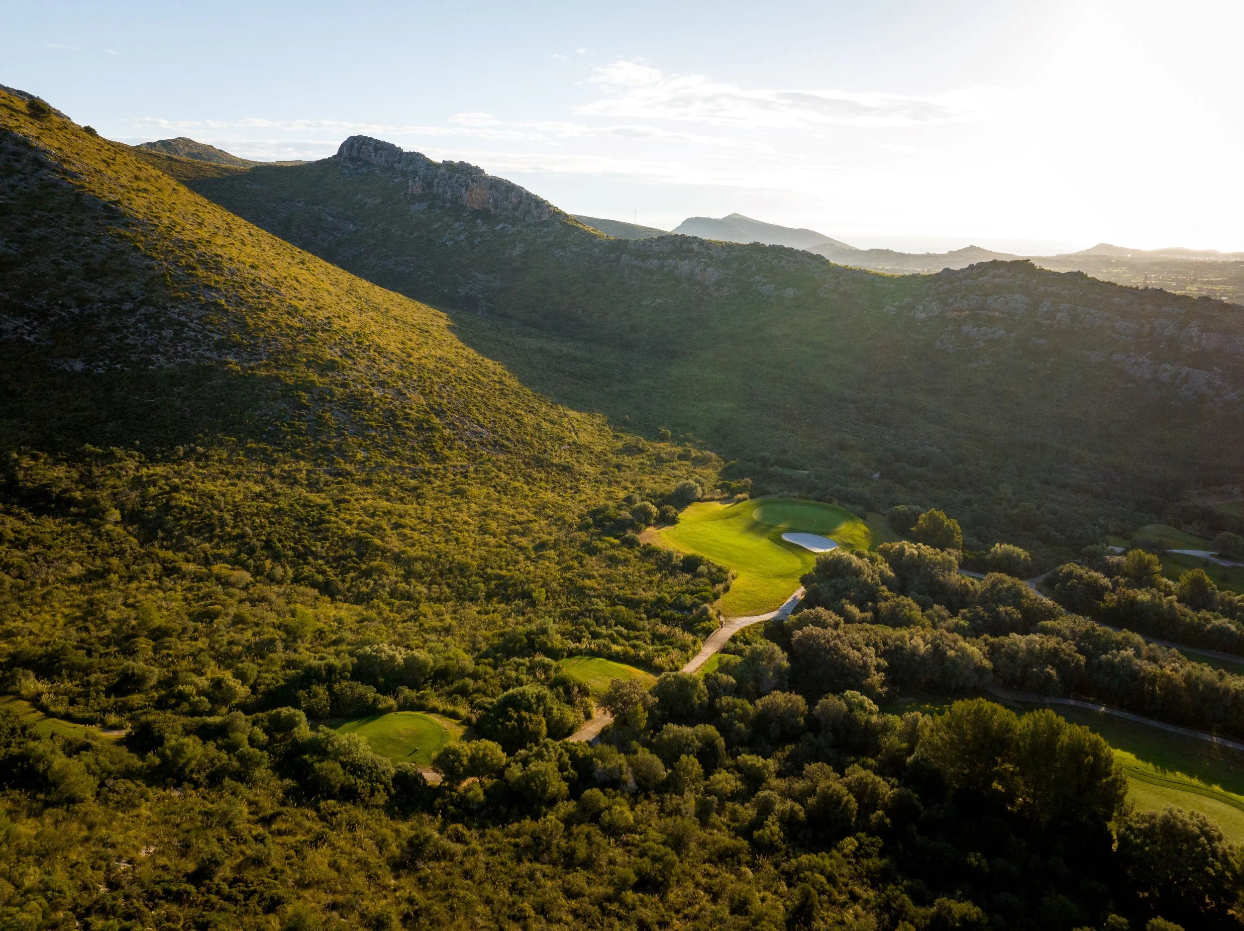Vista aérea de un campo de golf rodeado de colinas verdes y montañas al fondo, con senderos y áreas de césped bien cuidado.