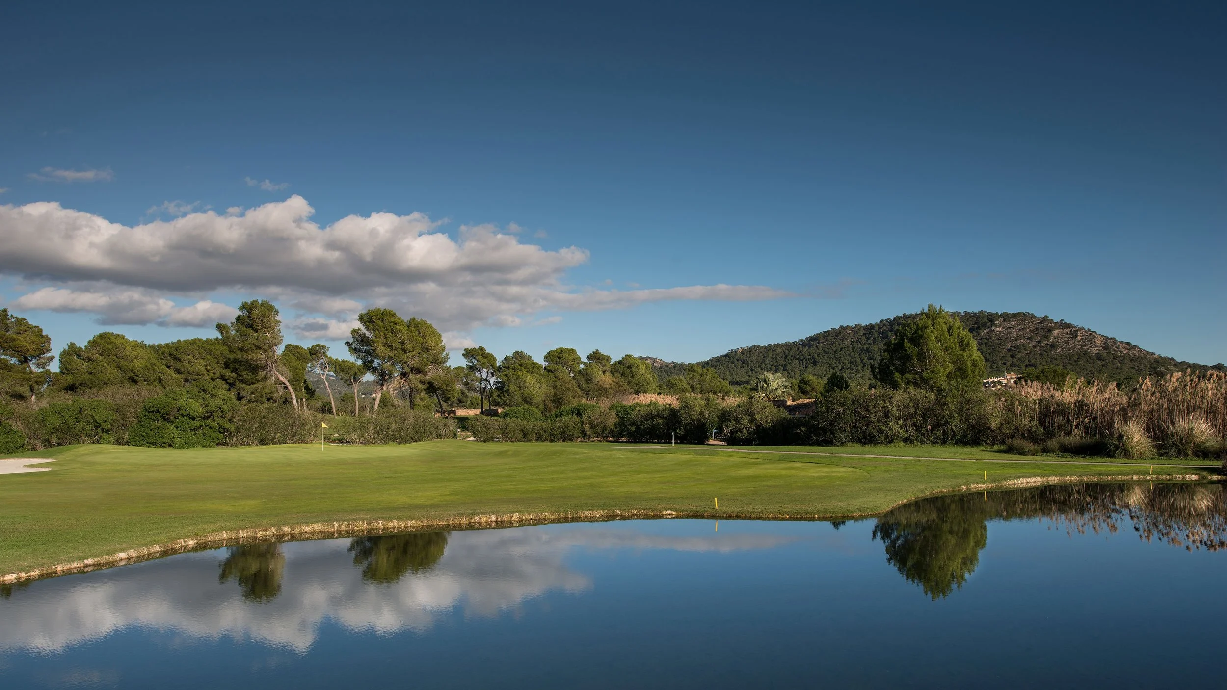 Campo de golf con árboles y montañas al fondo, cielo despejado con algunas nubes y reflejo en un estanque.
