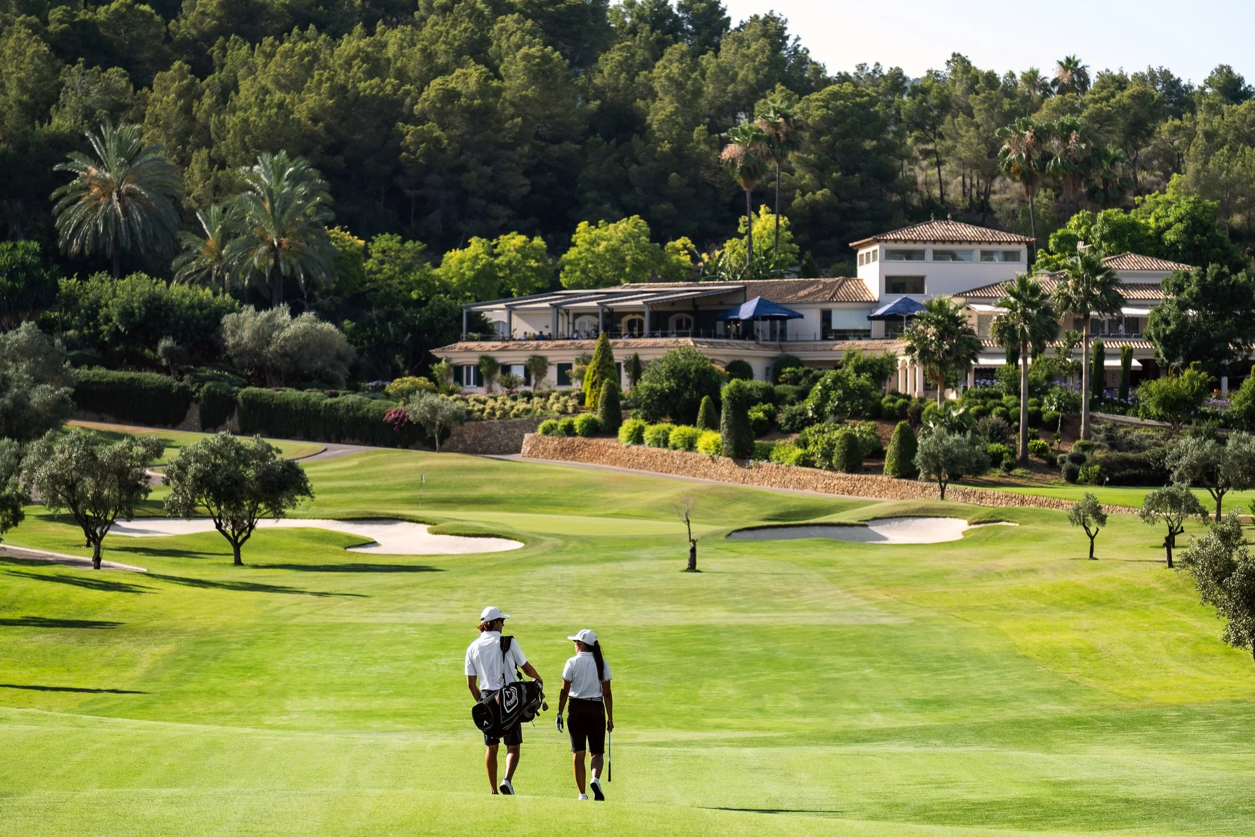 Dos golfistas caminan por un campo de golf verde con árboles, arroyos y un restaurante elegante en el fondo, rodeado de vegetación y palmeras.