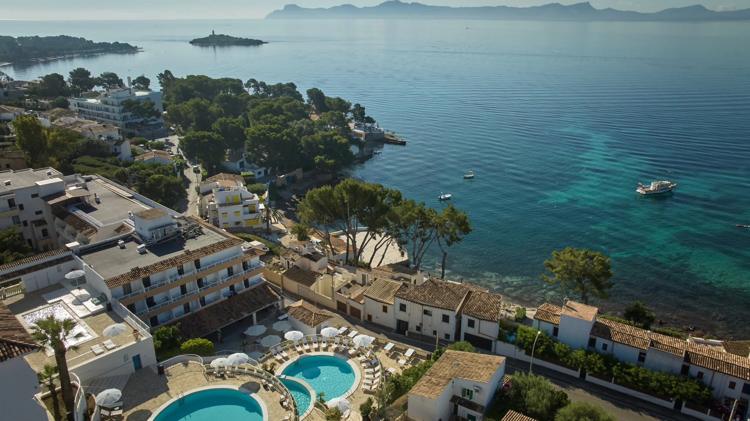 Vista aérea de un complejo hotelero junto al mar con piscinas, casas y árboles.
