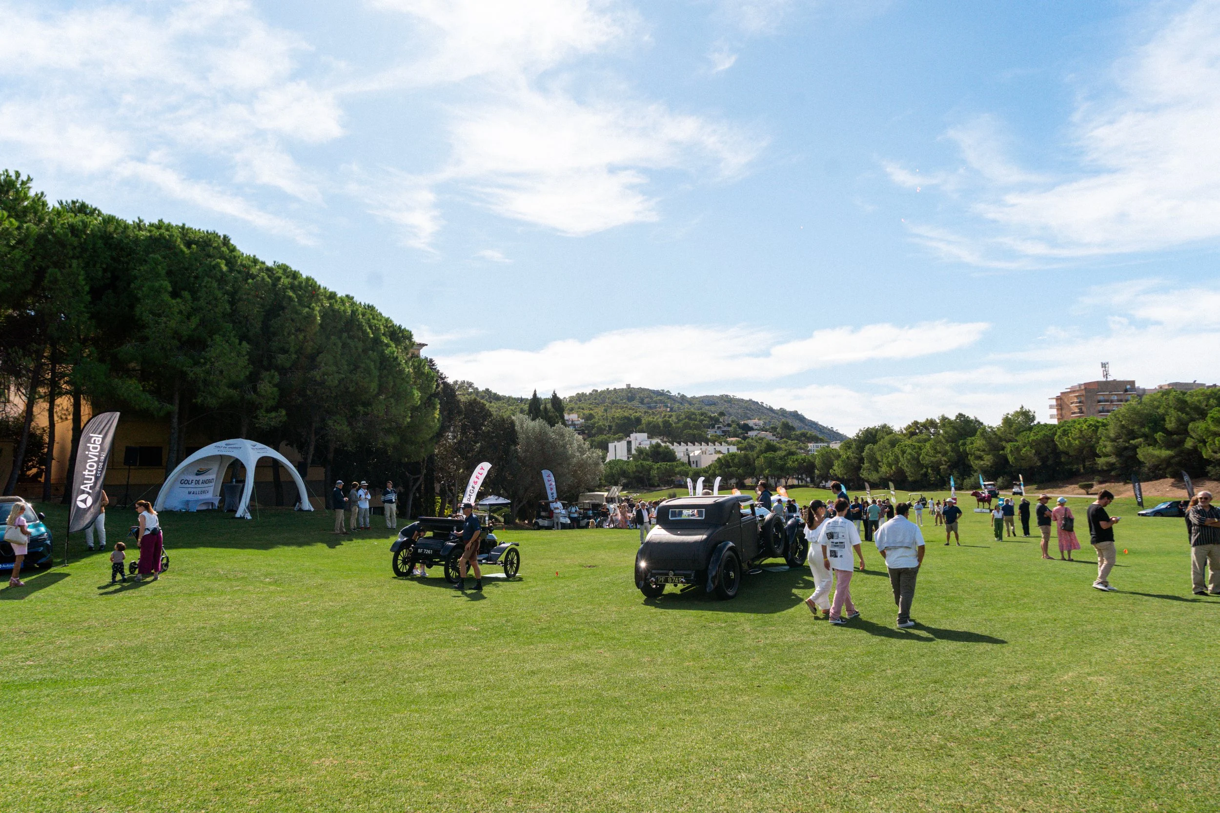 Un evento en un campo de césped con varias personas observando autos antiguos y modernos, banderas, y una estructura inflable blanca con logotipo de Autofin.