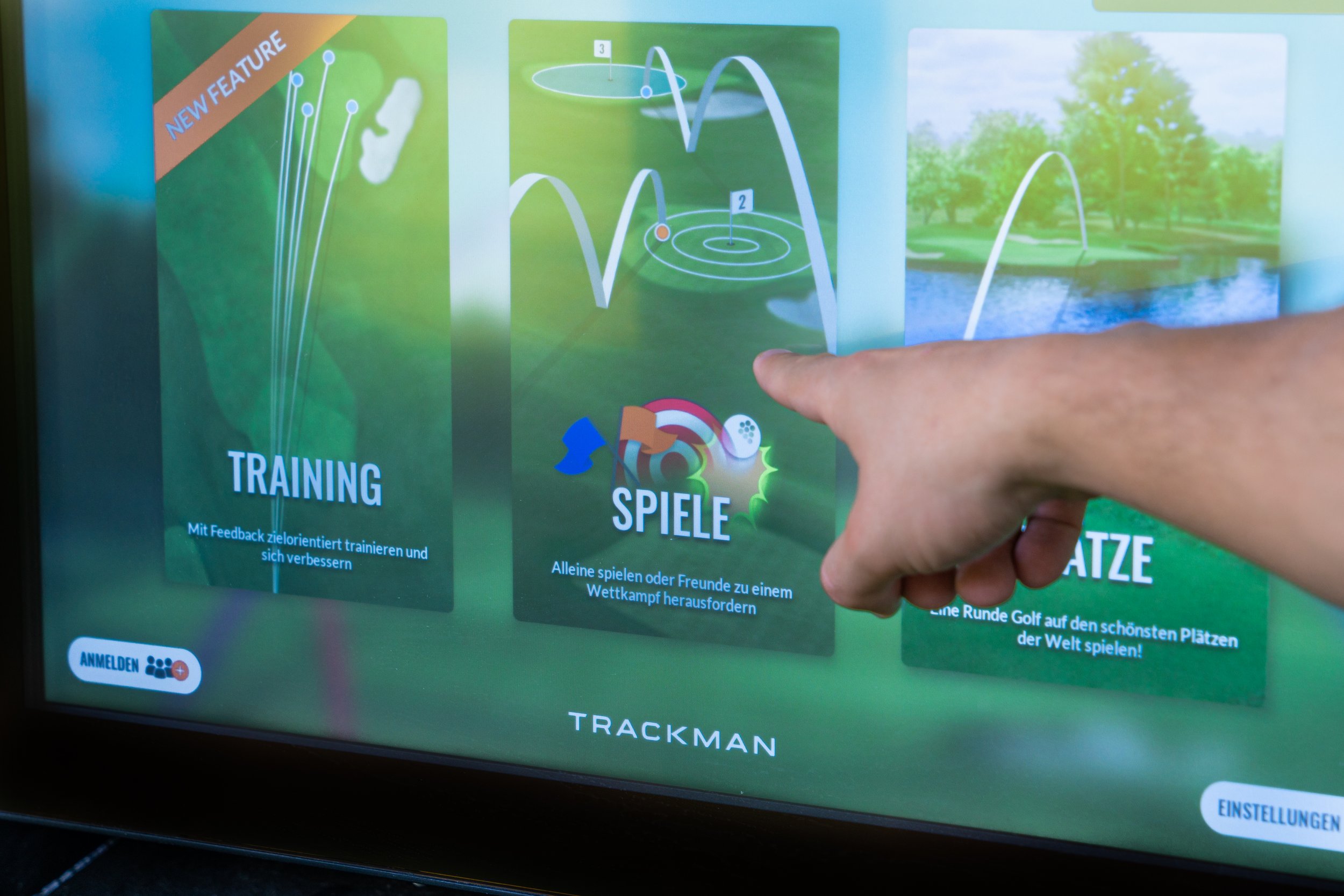 Pantalla táctil de una máquina de golf con opciones en alemán, incluyendo entrenamiento, juegos y jugar en los campos más hermosos del mundo, con una mano tocando la opción de 'Juegos'.