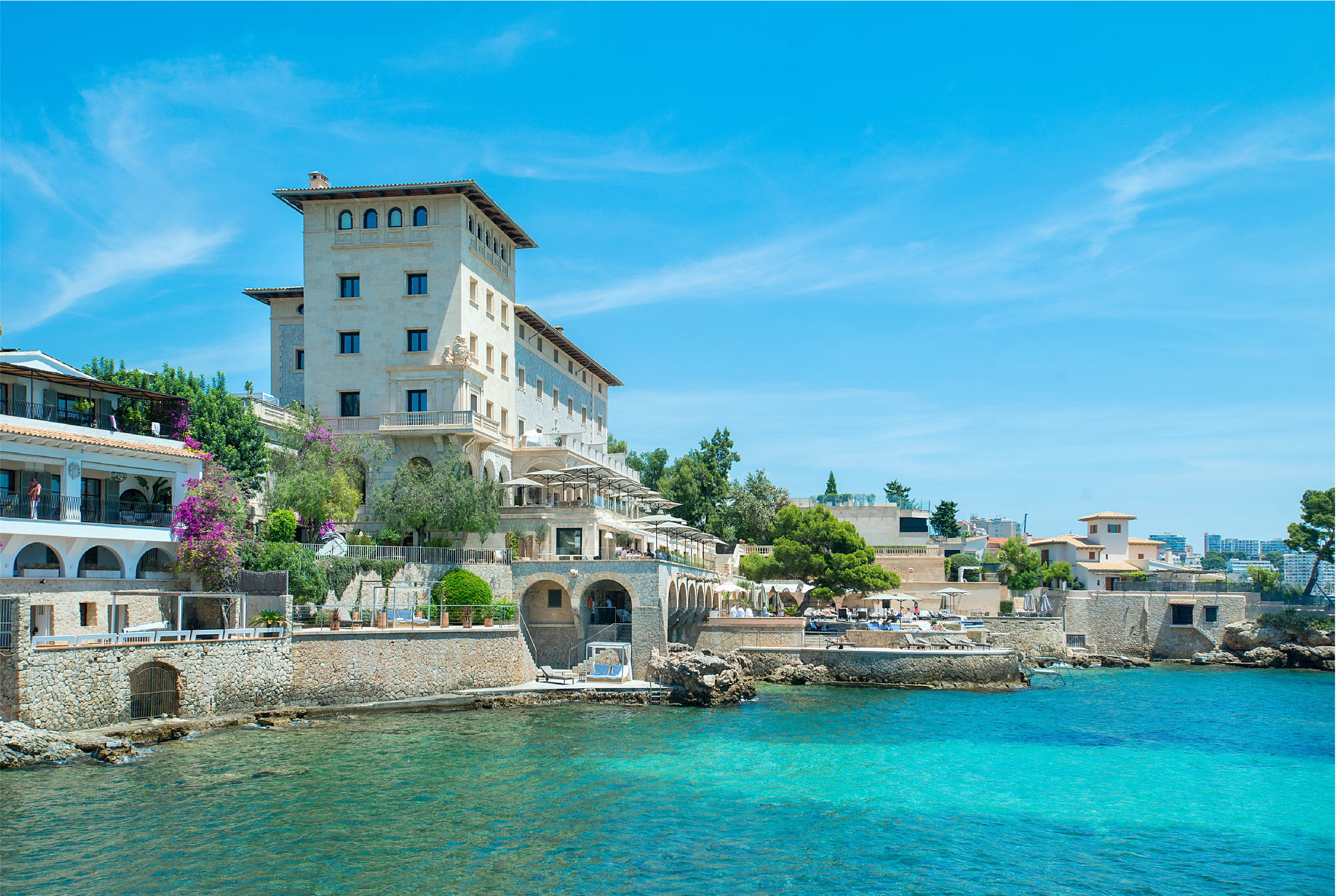 Edificio elegante junto al mar con balcones y vegetación, bajo un cielo azul claro.