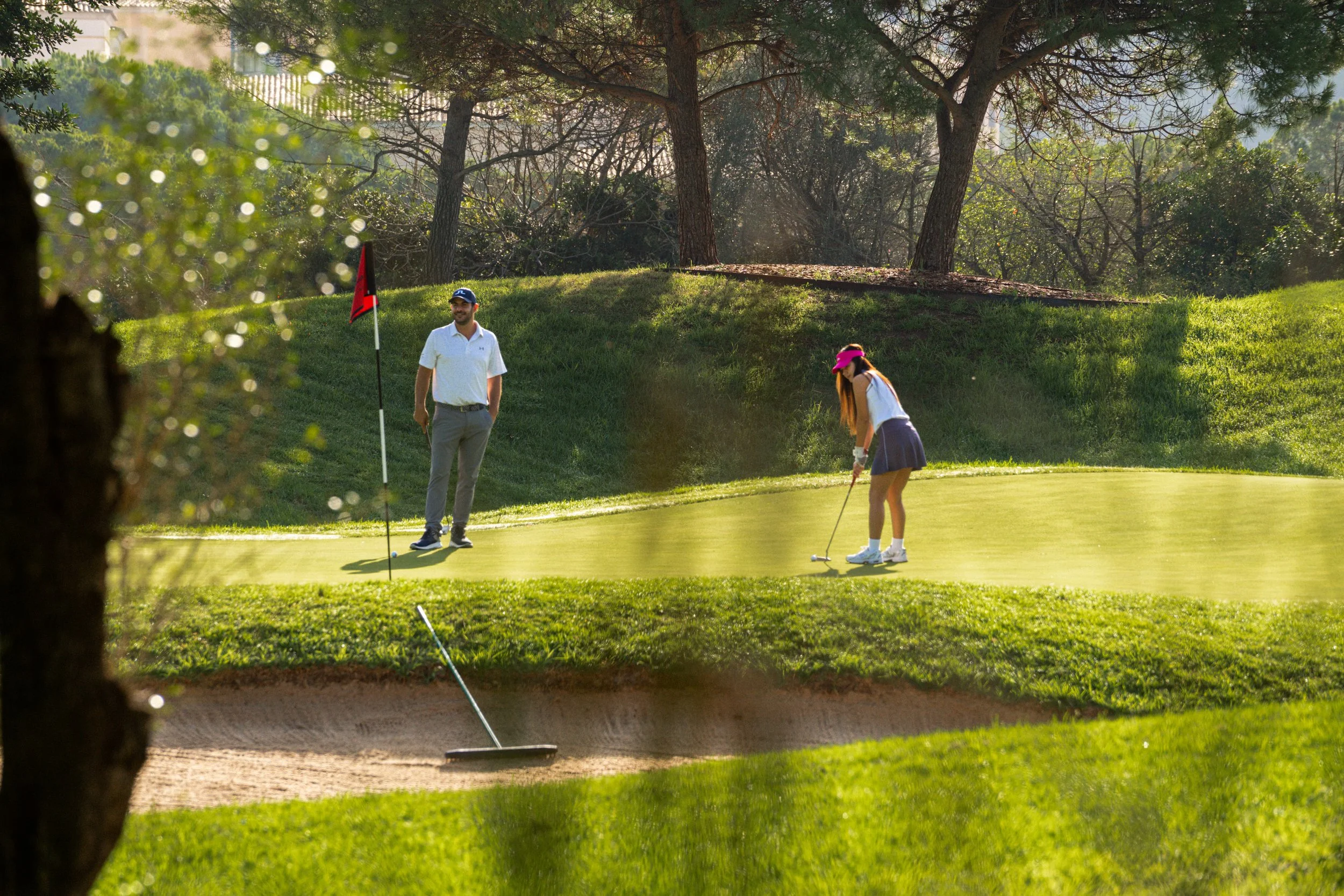 Dos personas jugando golf en un campo, una mujer está alineada preparando para golpear la pelota y un hombre observa, con árboles en el fondo.
