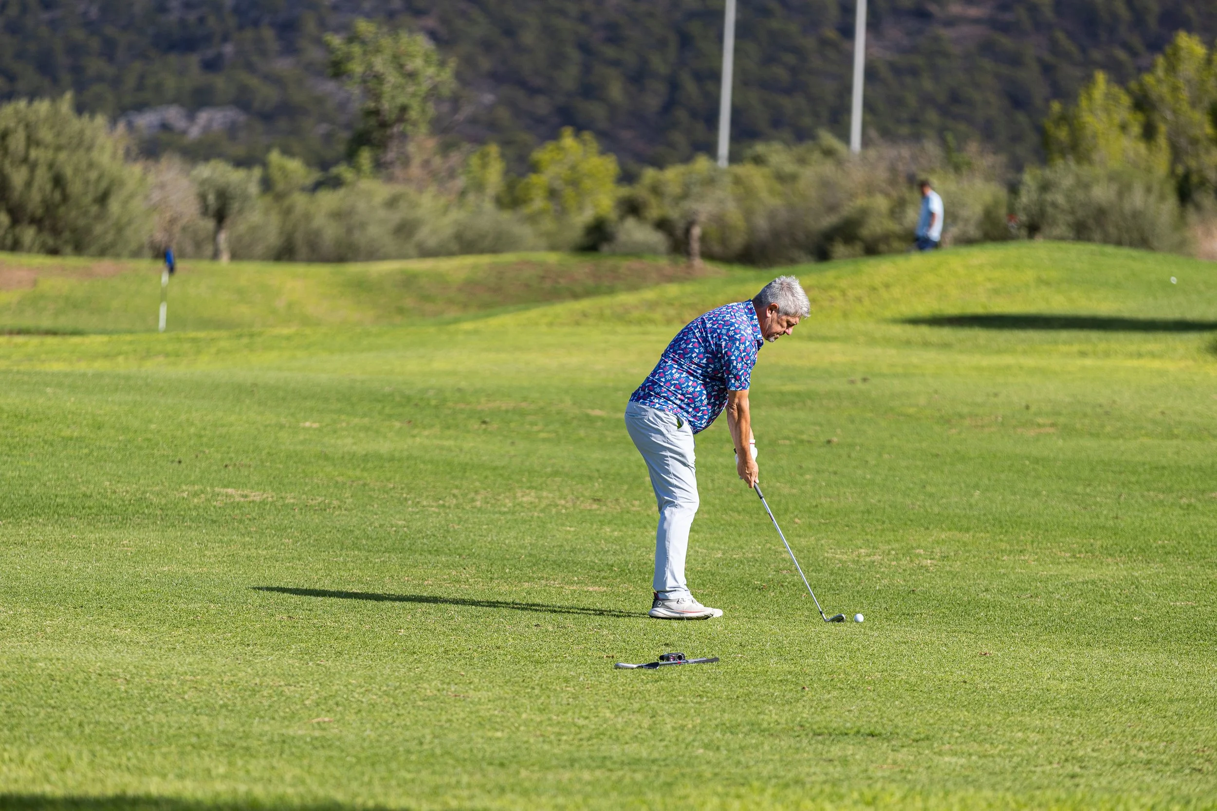 Un hombre mayor jugando golf en un campo verde con fondo de árboles y colinas.