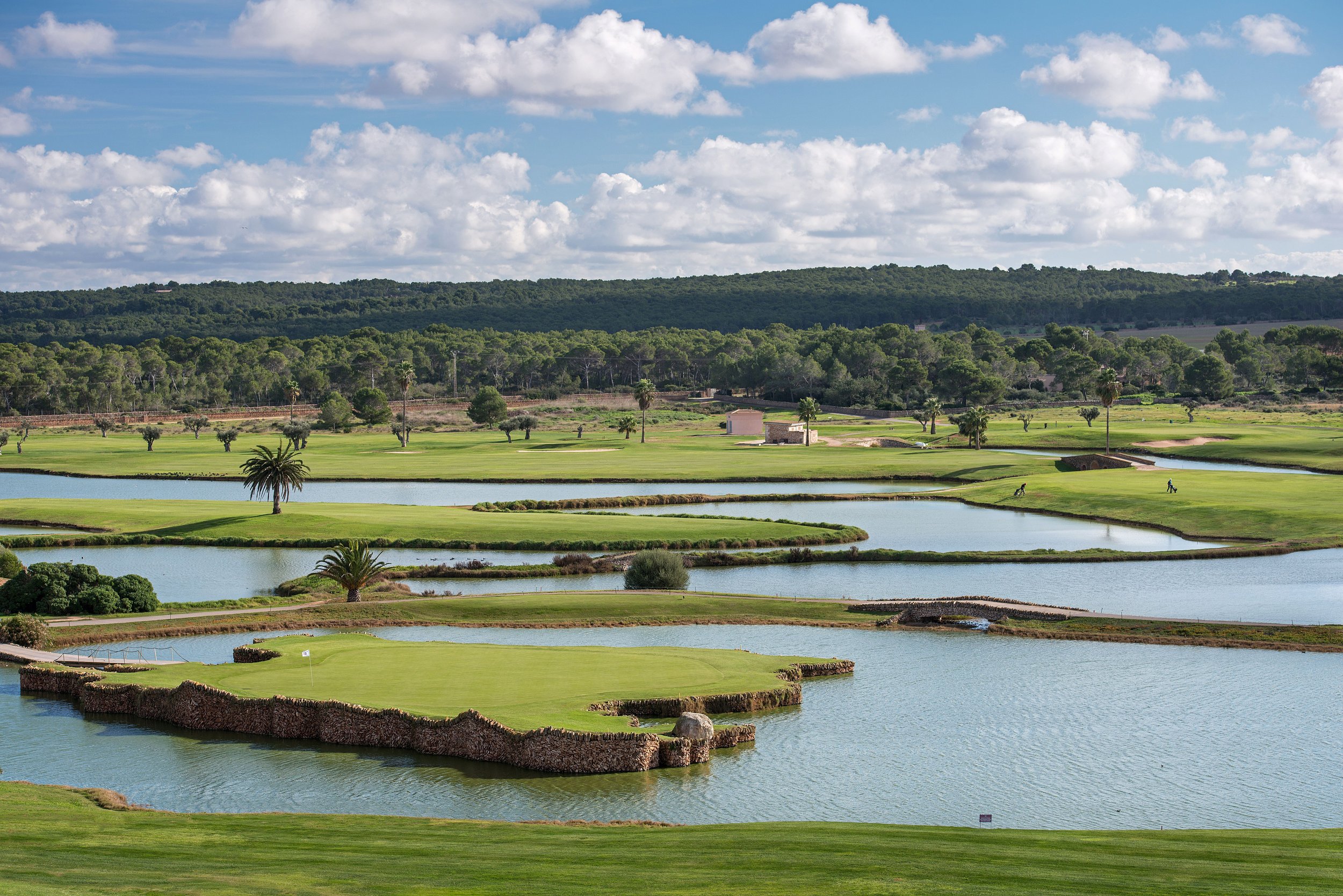 Campo de golf con lagos, árboles y colinas verdes bajo un cielo con nubes blancas y dispersas.