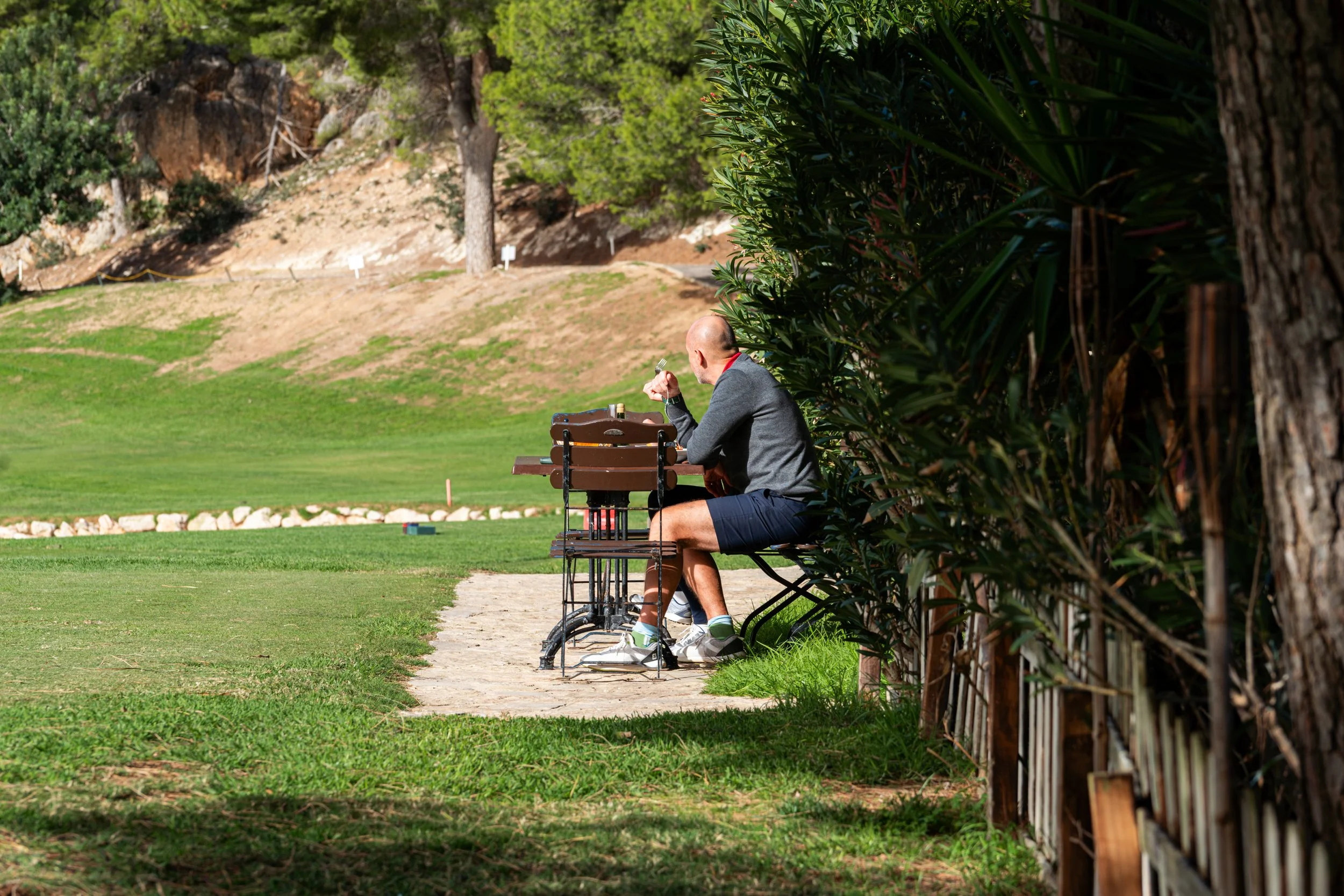 Un hombre sentado en un banco en un parque, junto a una mesa con sillas, disfrutando del día en un entorno verde con árboles y césped