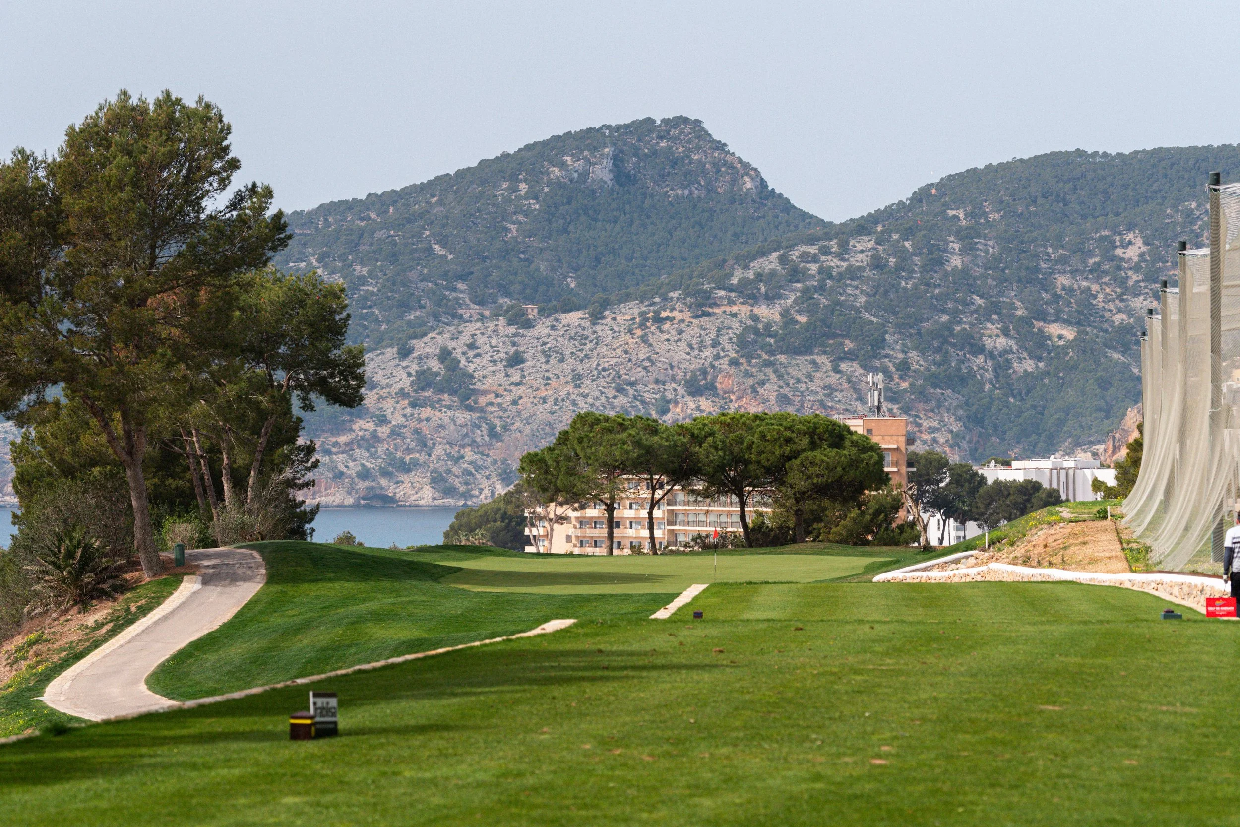 Un campo de golf con césped bien cuidado, rodeado de árboles y montañas en el fondo, y una parte de un edificio visible al fondo.