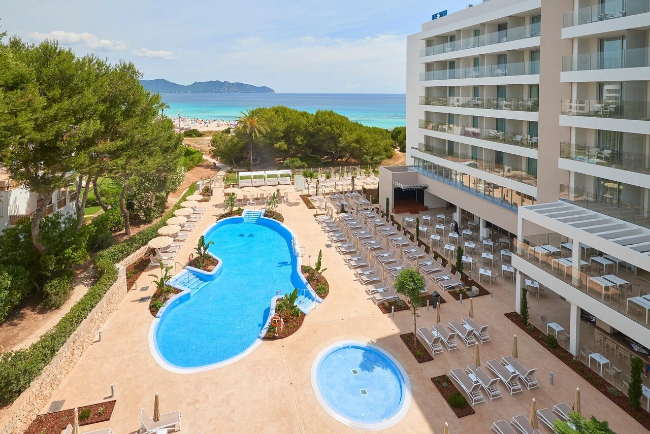 Vista del complejo hotelero con piscina y playa en el fondo, edificio de habitaciones a la derecha y área de descanso con sillas y sombrillas.