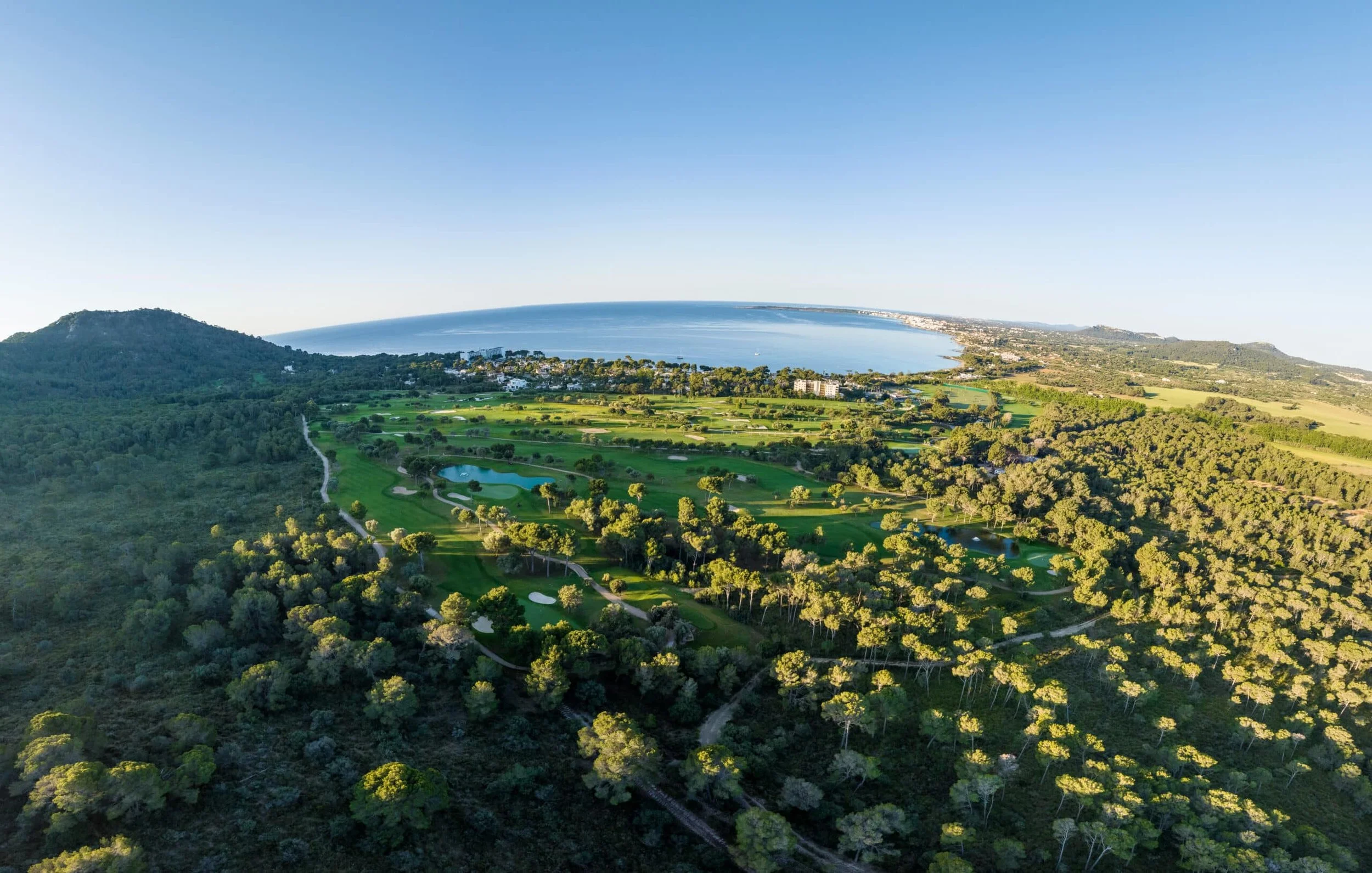 Vista aérea de un campo de golf junto a la costa, con áreas verdes, árboles y pequeños lagos, y el océano en el fondo bajo un cielo despejado.