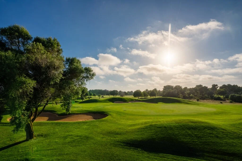 Campo de golf con árboles, arena y colinas verdes bajo cielo soleado con nubes