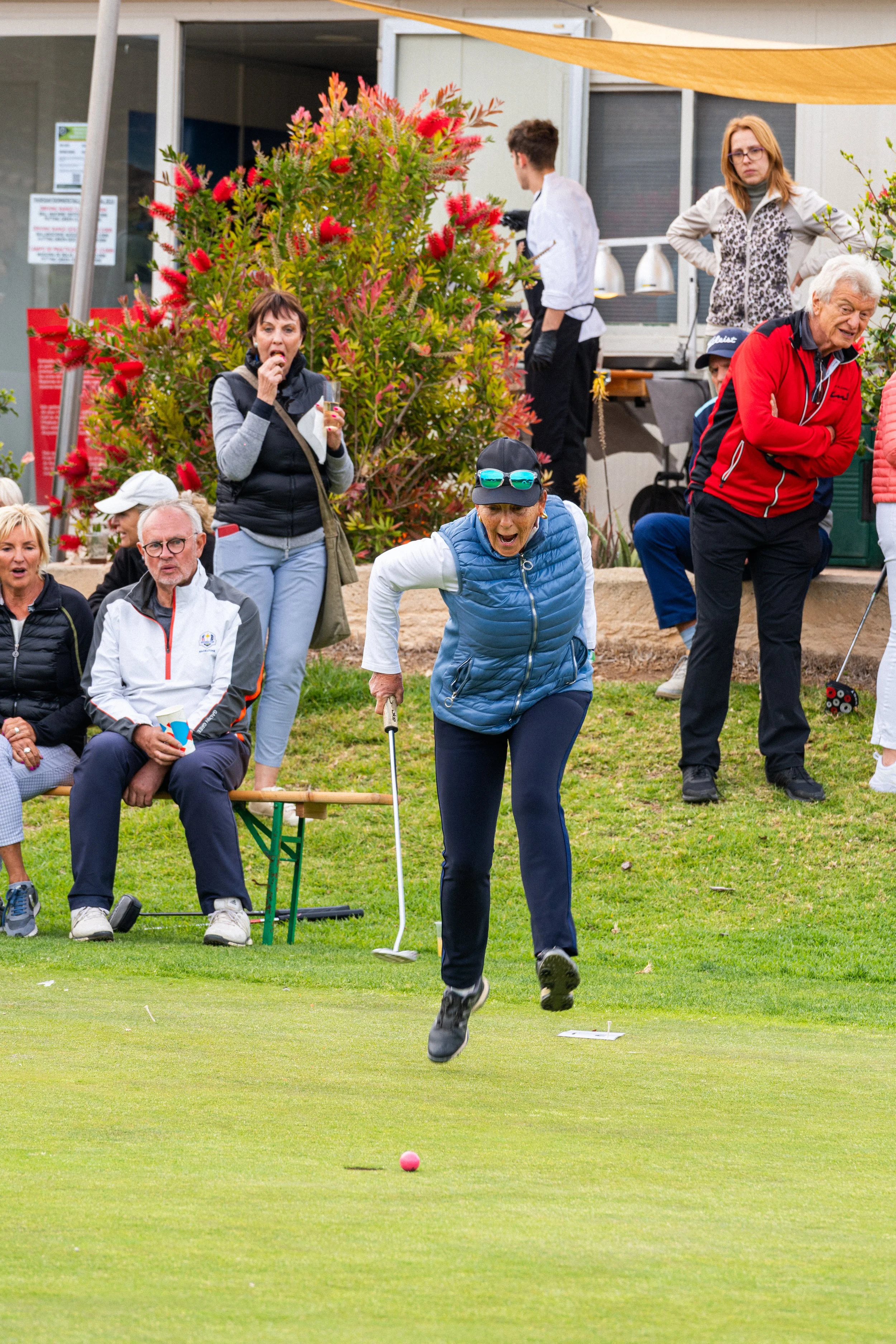 Mujer mayor jugando golf en un campo, rodeada de varias personas observando y disfrutando del juego.