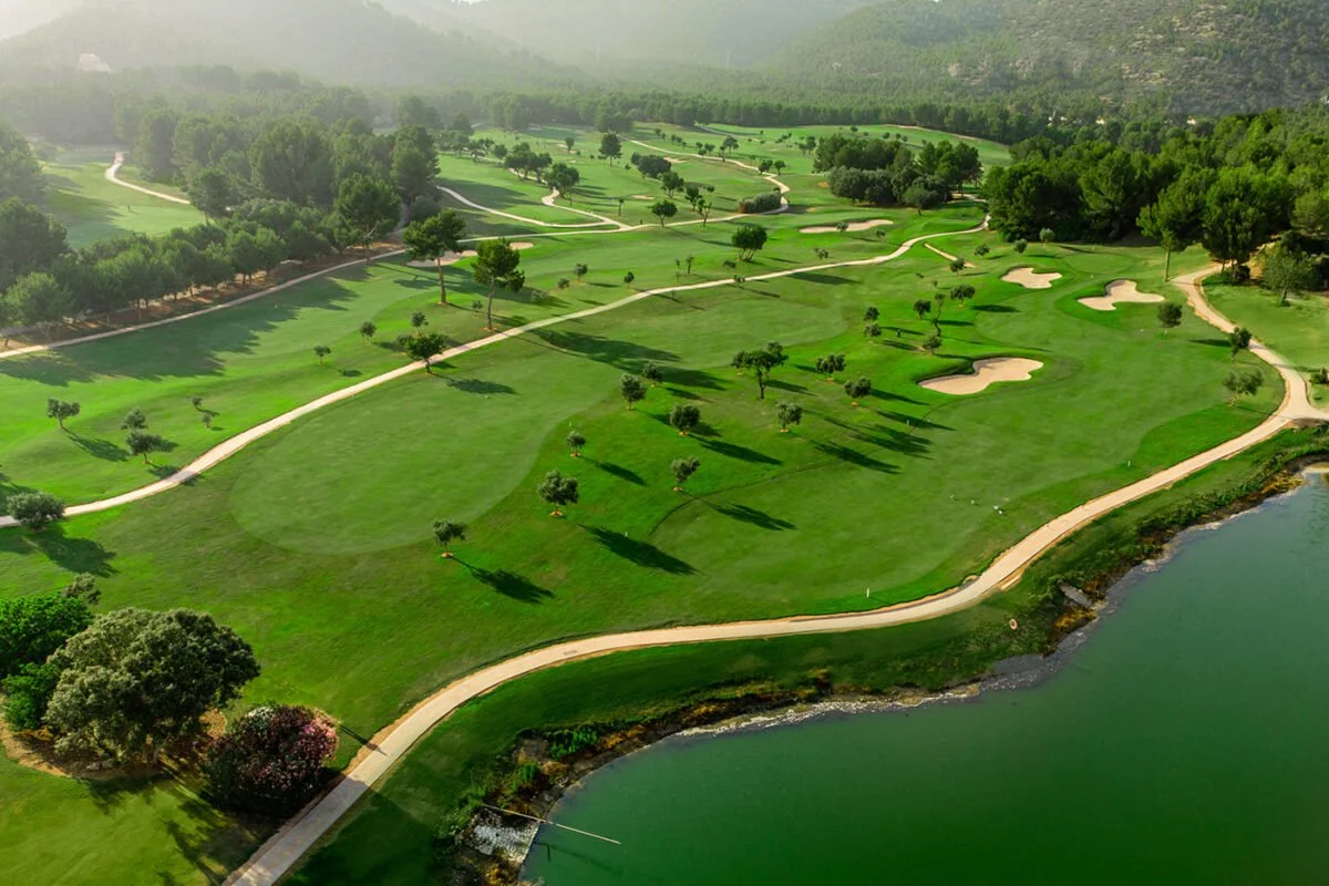 Campo de golf con áreas verdes, árboles, caminos y bunkers junto a un cuerpo de agua con montañas al fondo