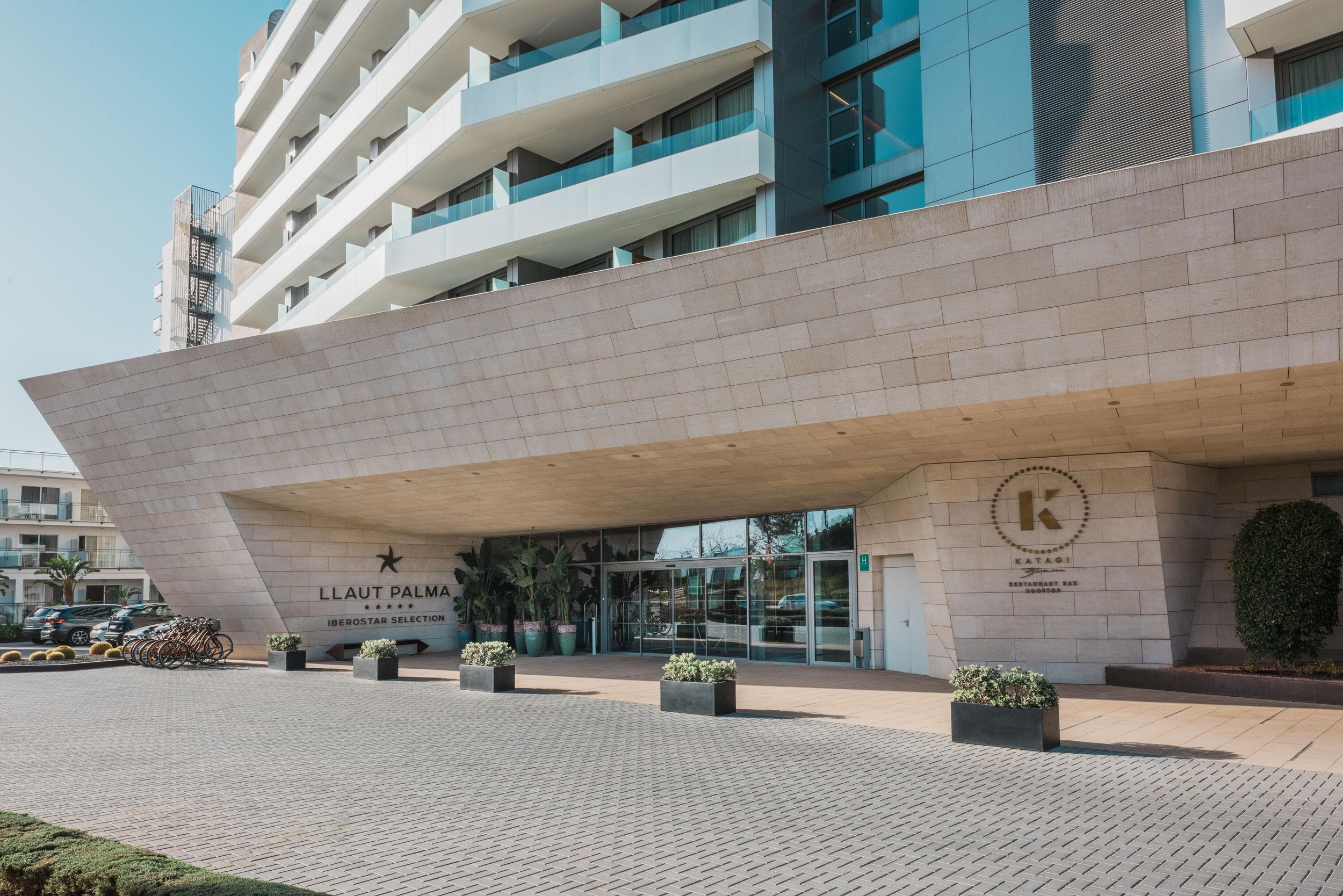 Fachada del edificio del hotel Llaut Palma en la ciudad, con entrada principal, plantas en macetas y área de estacionamiento con coches y bicicletas.