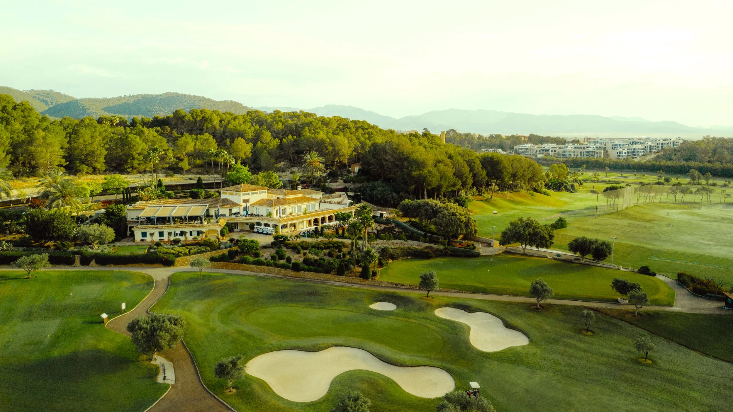 Vista panorámica de un campo de golf con áreas verdes, búnkeres y caminos, rodeado de árboles y colinas en el fondo.