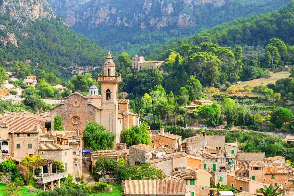Una vista panorámica de un pueblo en una zona montañosa con casas de techos de tejas, una iglesia con campanario y muchas áreas verdes con árboles y arbustos.