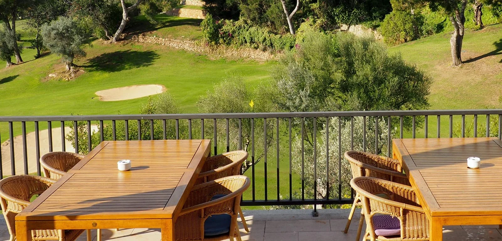 Dos mesas de madera con sillas de mimbre en una terraza, con vista a un campo de golf con césped, árboles y una pequeña barda de piedra.