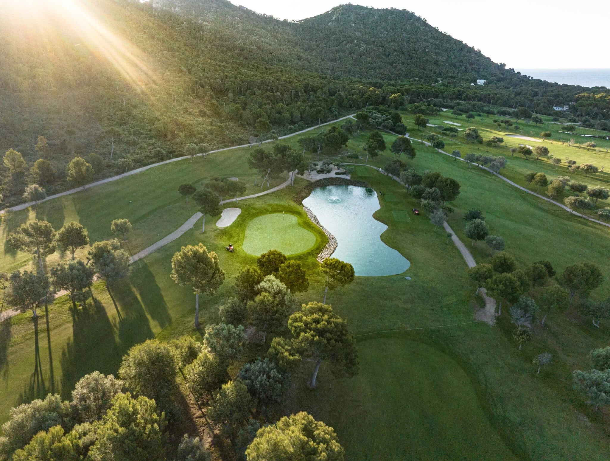 Vista aérea de un campo de golf con varios lagos y árboles, en un área montañosa con el sol brillando desde la izquierda.