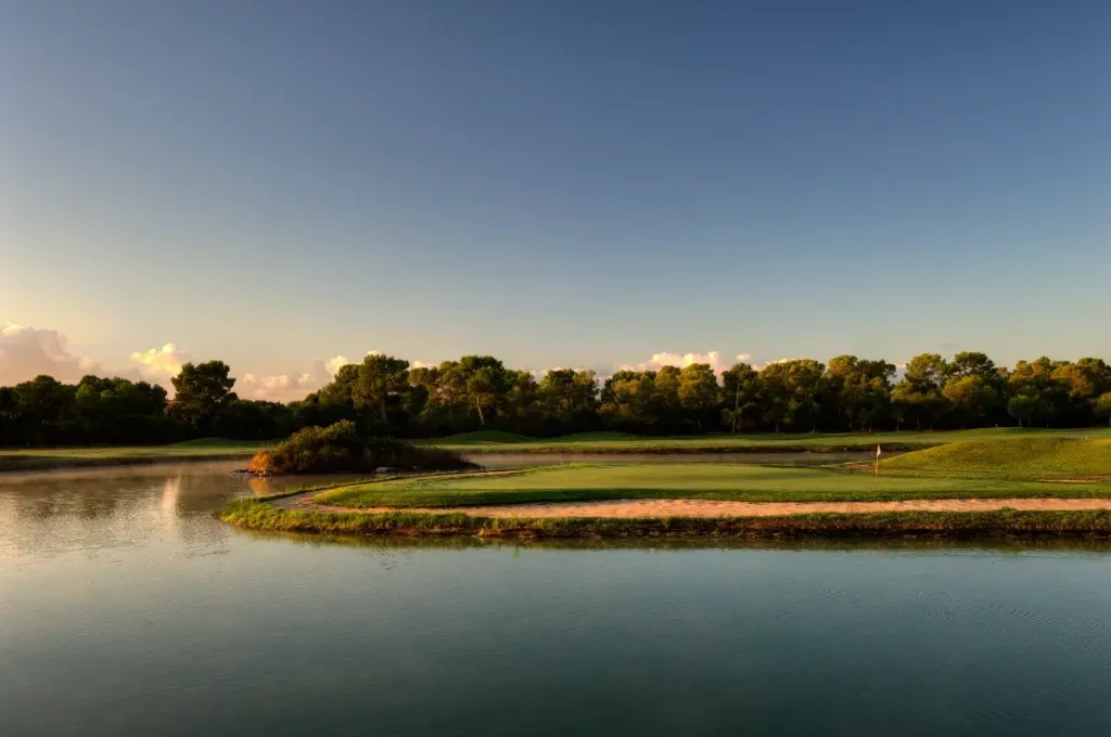 Vista de un campo de golf al atardecer, con un lago en primer plano y árboles en el fondo.