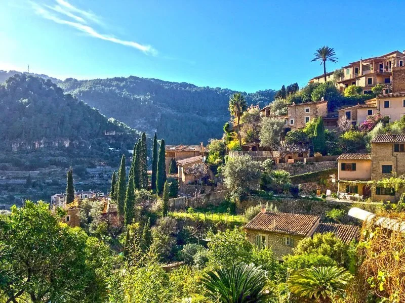 Paisaje de colinas con casas y vegetación verde, incluyendo árboles y palmeras, bajo un cielo azul con algunas nubes.