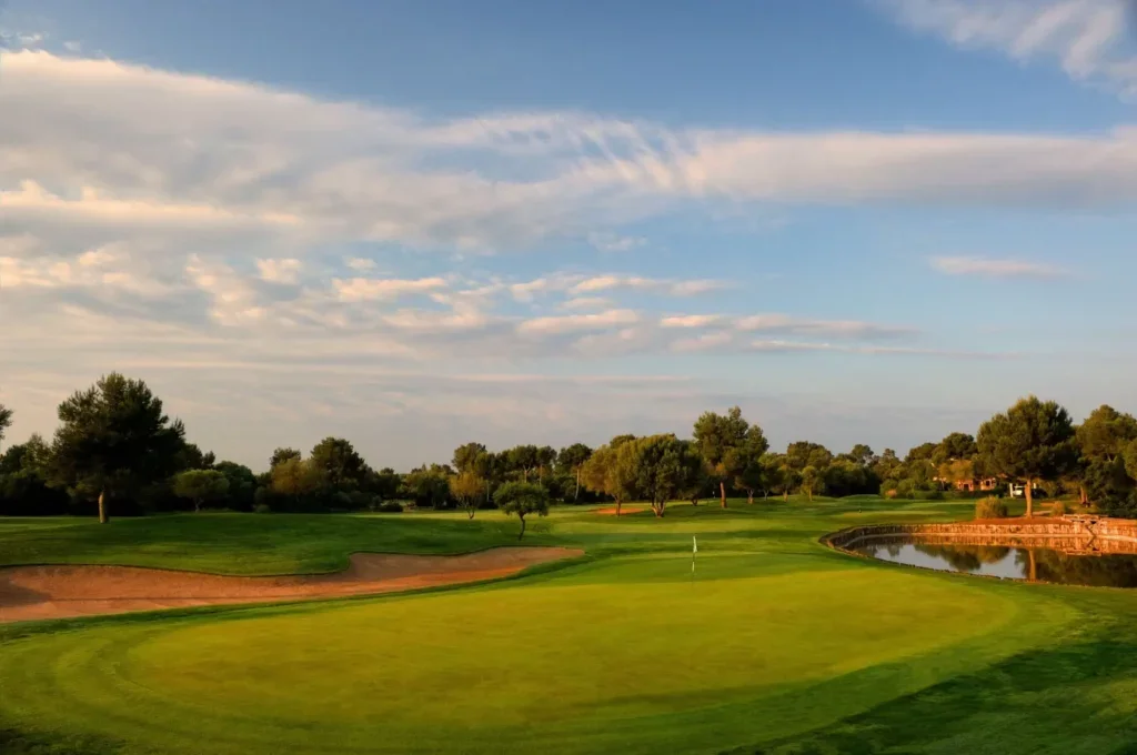 Campo de golf con césped bien cuidado, árboles dispersos, un estanque y cielo despejado con nubes delgadas.
