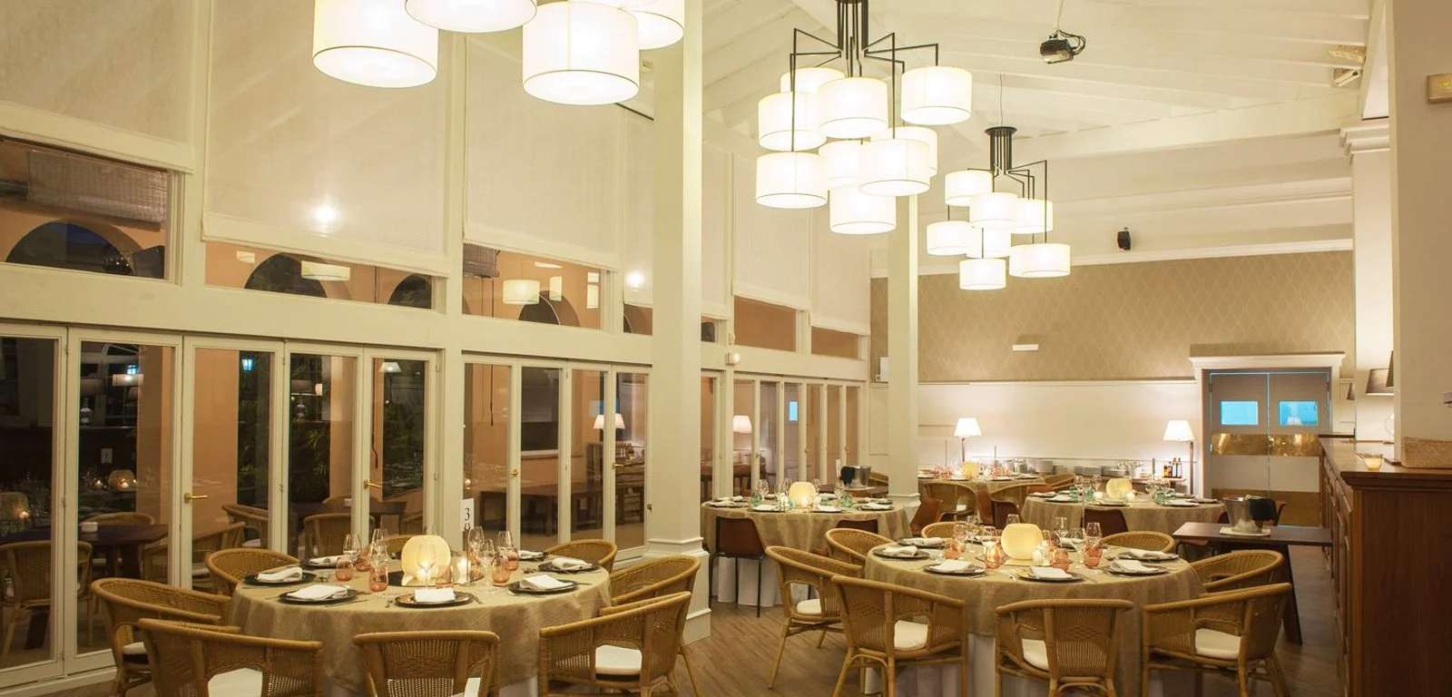 Interior de un restaurante elegante con mesas y sillas de mimbre, iluminación cálida y paredes decoradas, preparado para cenas con manteles, copas y velas.
