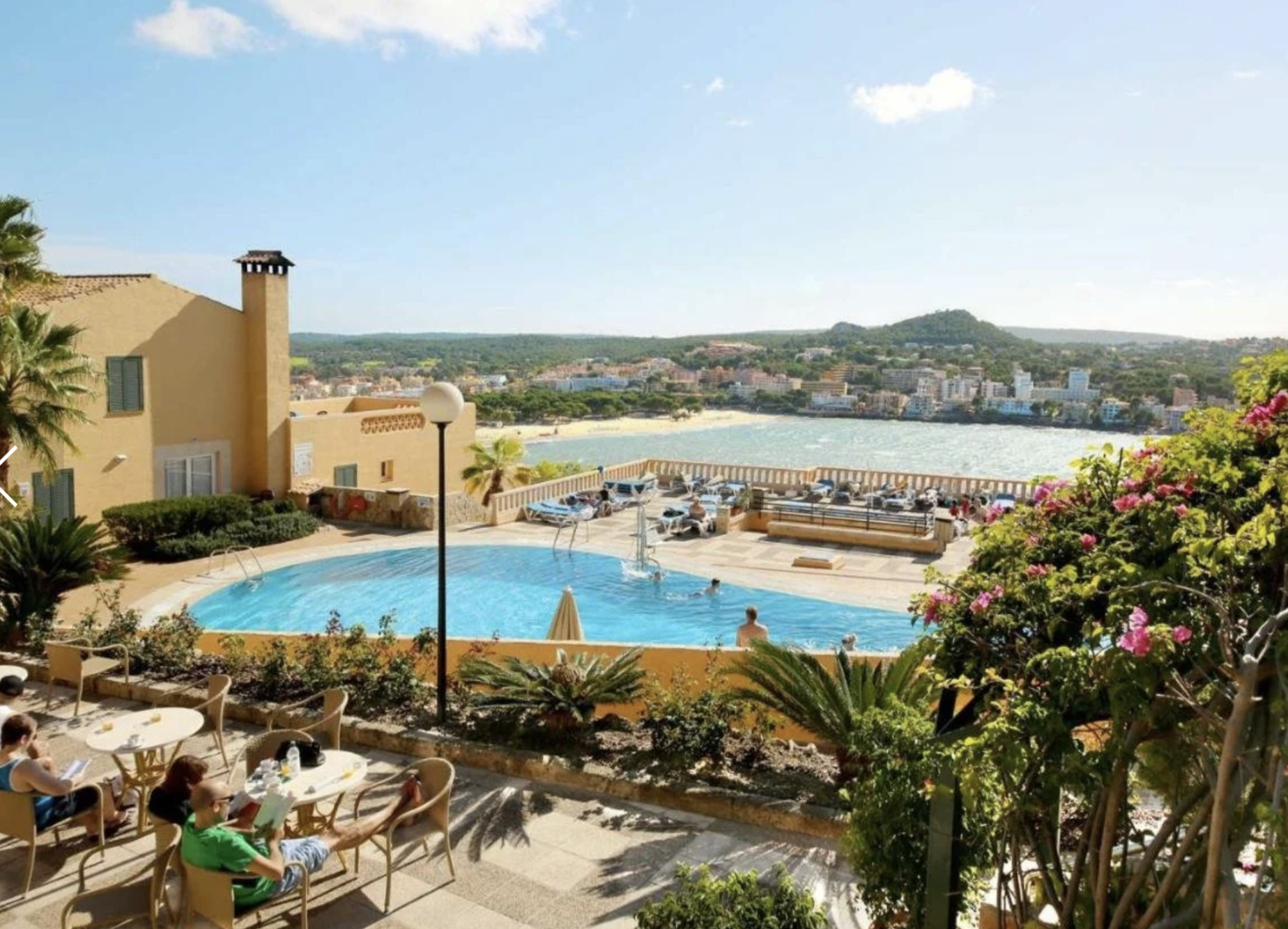 Vista de una piscina y un área de descanso con personas sentadas en sillas y mesas, un lago y una ciudad con montañas al fondo, en un día soleado.