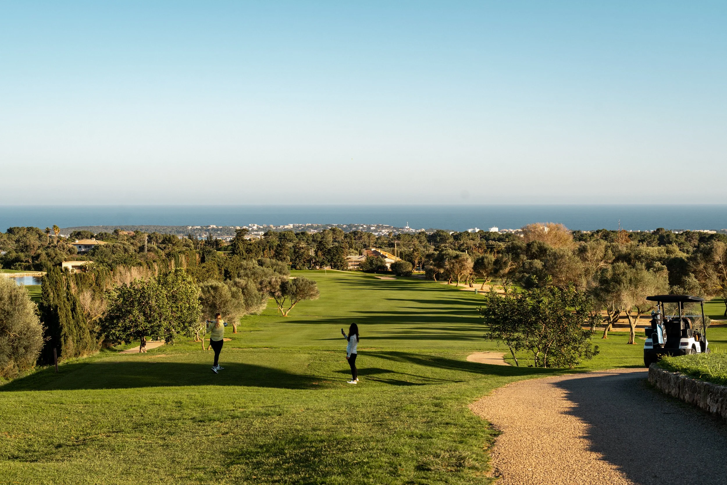Dos personas jugaron golf en un campo con vista al océano, rodeado de árboles y vegetación, y un carrito de golf a un lado.