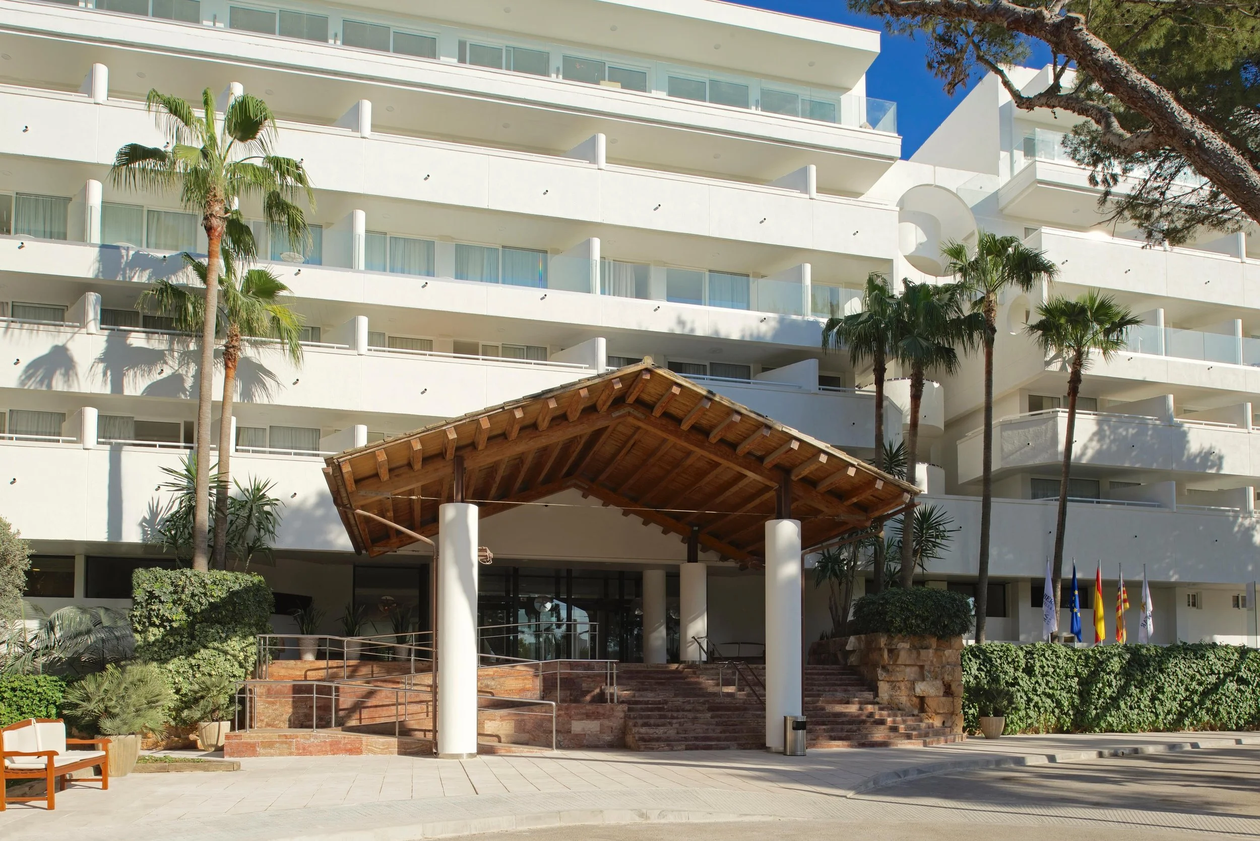 Edificio moderno de varios pisos de color blanco con balcones de cristal, palmeras y jardines en el exterior, bajo un cielo despejado.