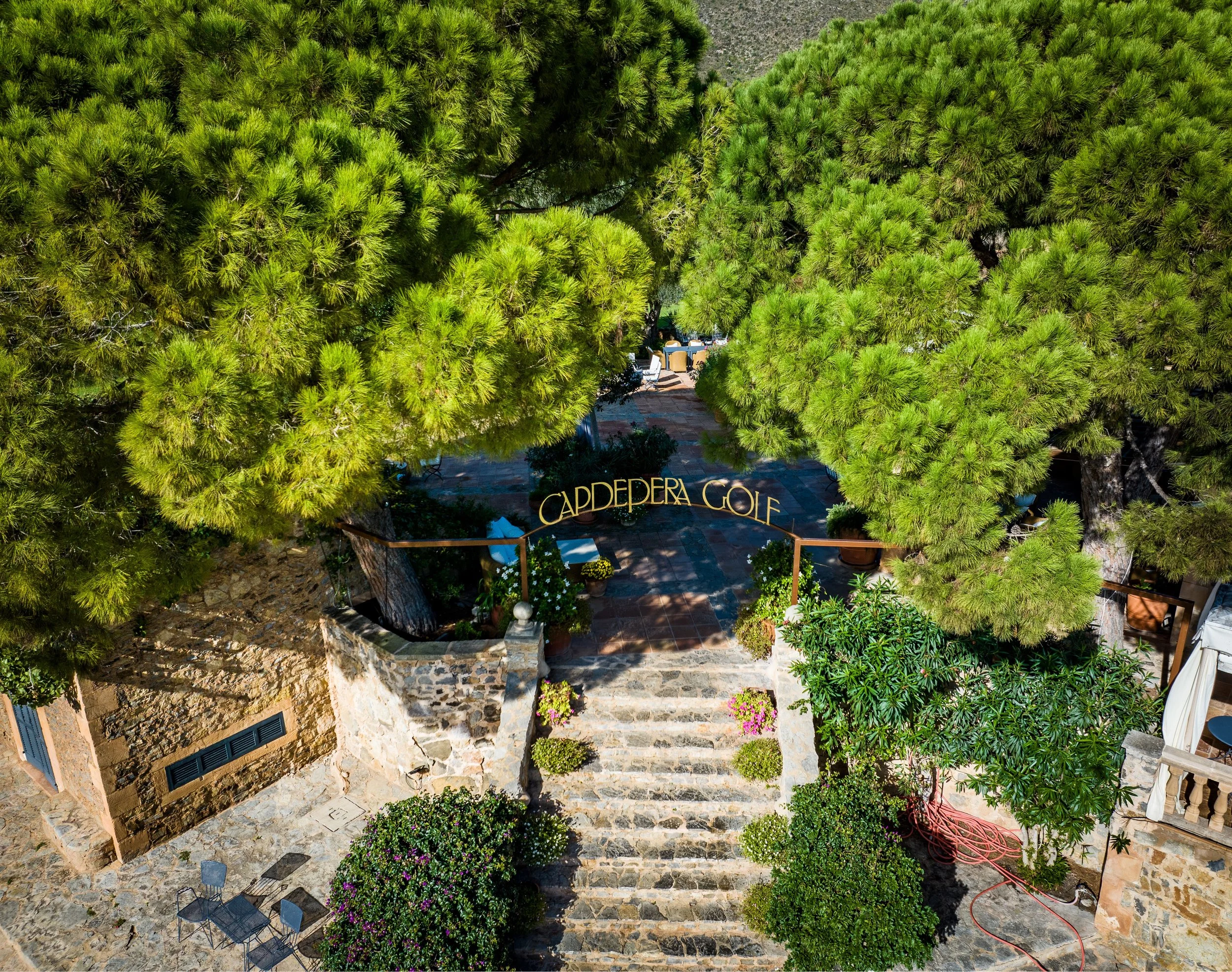 Vista aérea de un camino de piedra entre árboles grandes y verdes en un entorno de jardín con escaleras y áreas de césped decoradas con plantas y flores.