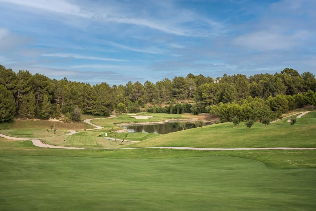 Paisaje de campo de golf con grama bien cuidado, caminos, árboles y laguna bajo cielo parcialmente nublado.