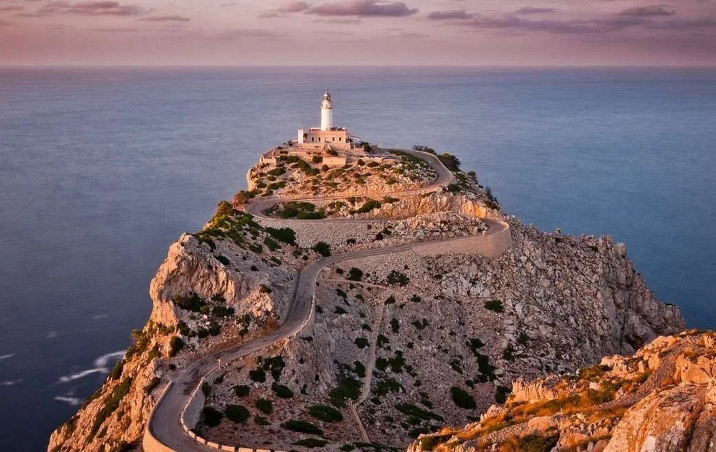 Lighthouse on a rocky island, rode winding up the hillside, al atardecer con cielo rosado y mar azul en el fondo.