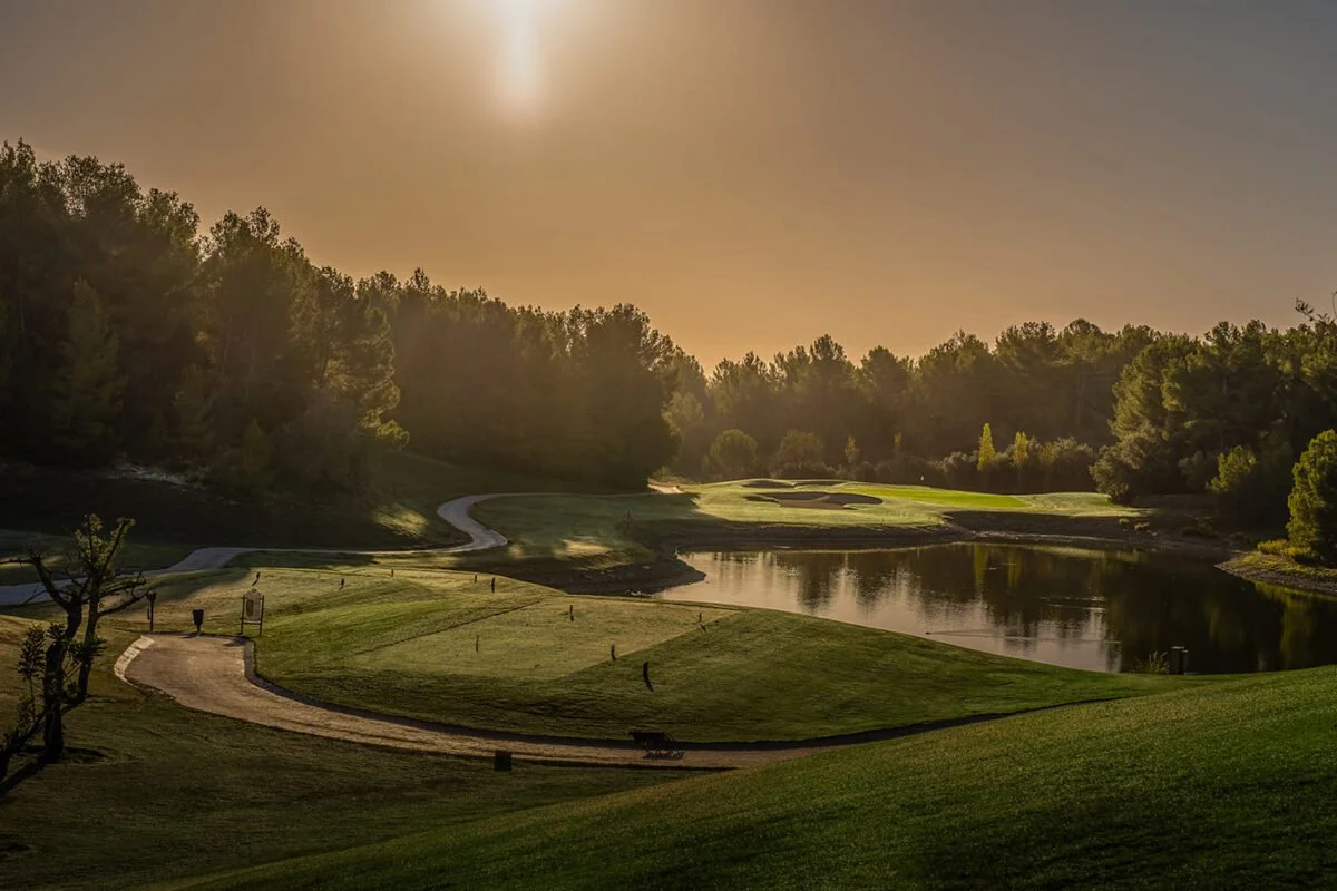 Paisaje de un campo de golf con árboles, agua y caminos, bajo un cielo soleado.