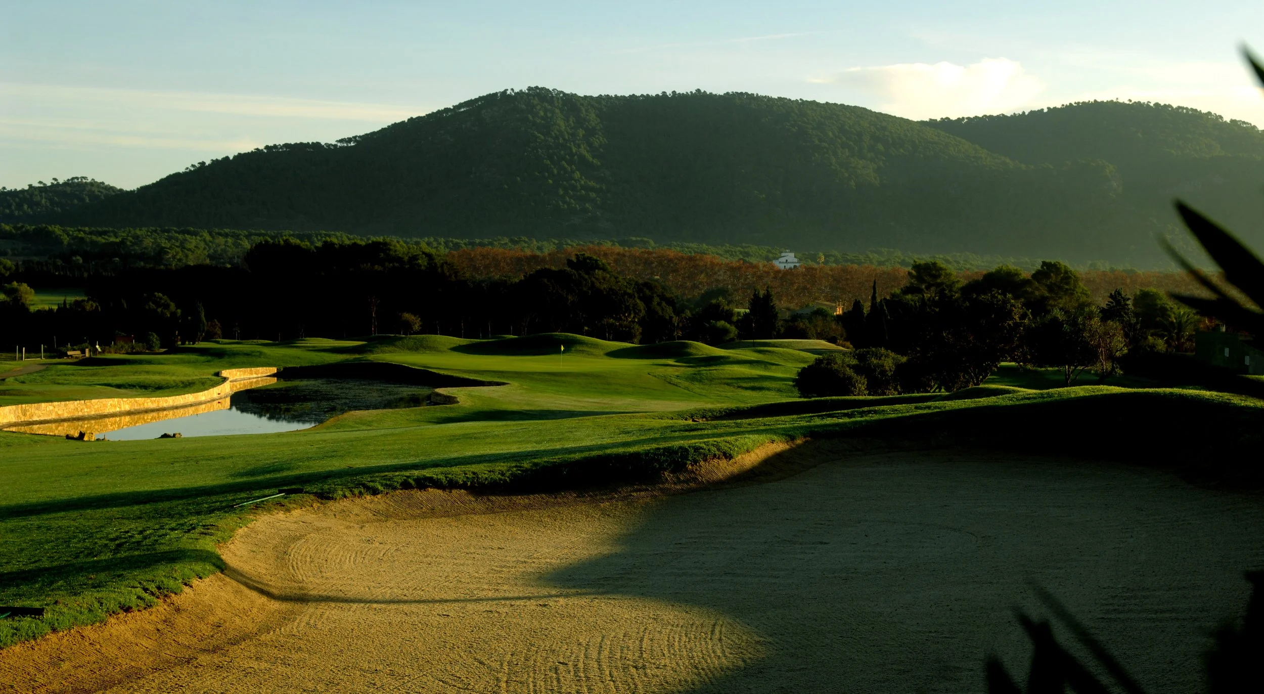 Campo de golf con vegetación y colinas al fondo, con un lago y un bunker en primer plano.