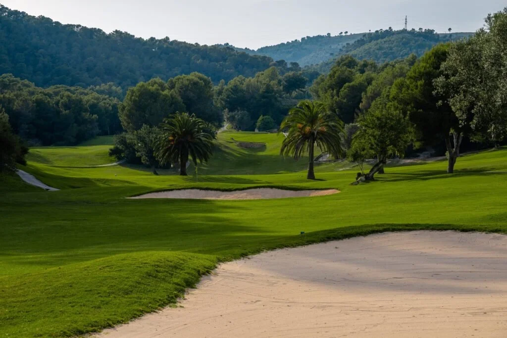 Campo de golf con arena y árboles verdes, rodeado de colinas y montañas.
