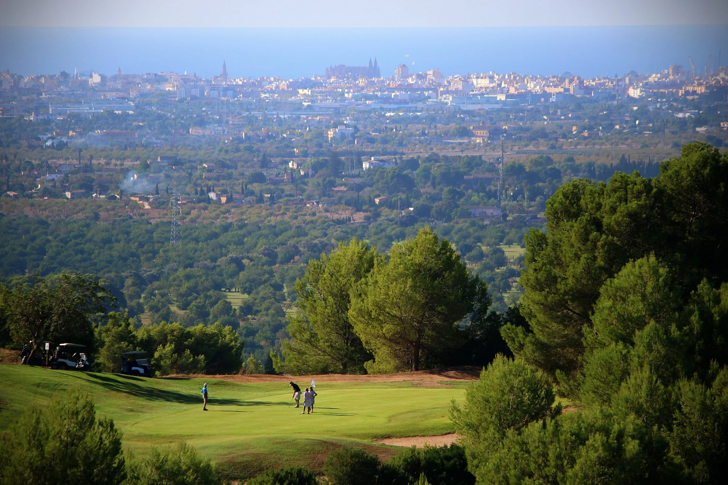 Vista de un campo de golf con jugadores, rodeado de árboles verdes y ciudad en el fondo con edificios y el mar