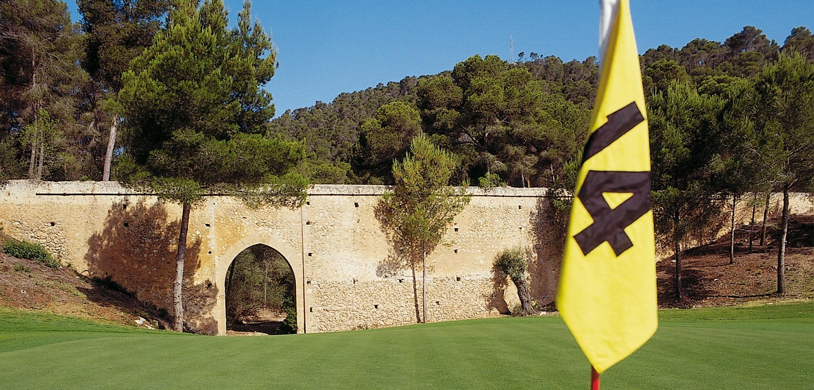 Vista de un campo de golf con bandera amarilla y una estructura de piedra con un arco, rodeada de árboles verdes y colinas, bajo un cielo azul.