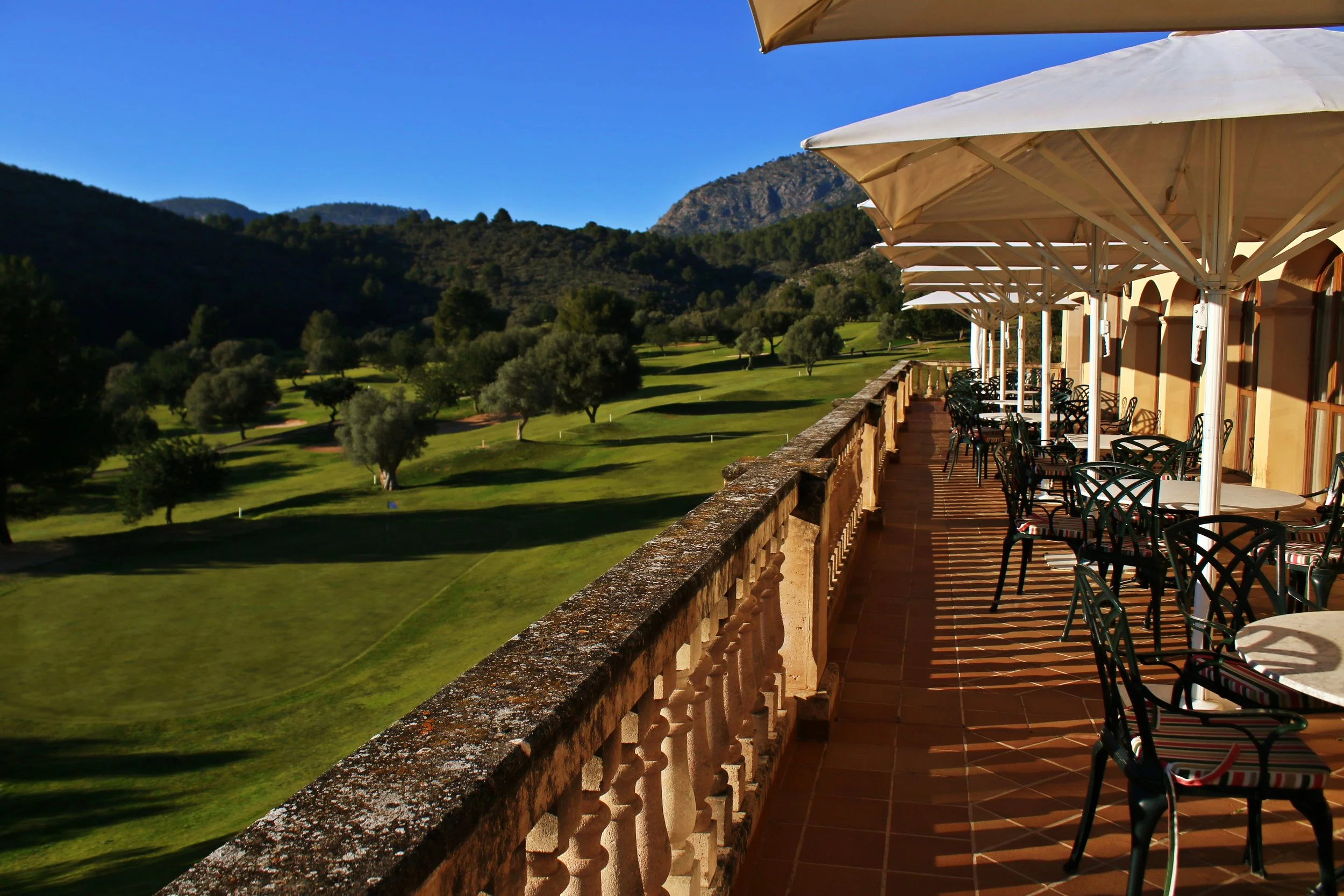 Terraza con mesas y sillas, vista a un campo de golf con árboles y montañas al fondo, en un día soleado