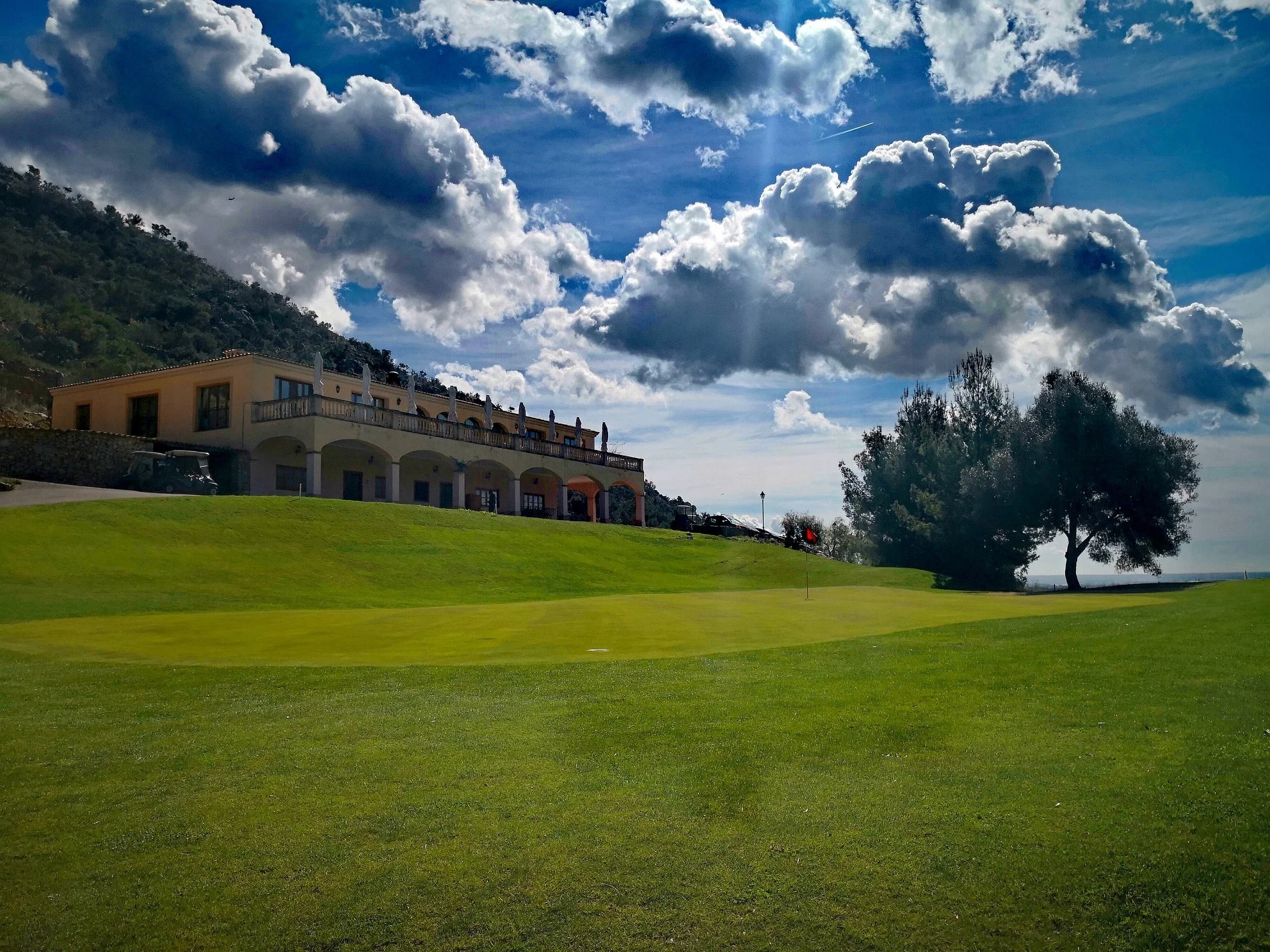 Vista del campo de golf con césped bien cuidado, un edificio con terraza en la parte superior y árboles, con un cielo nublado y sol brillando entre las nubes.