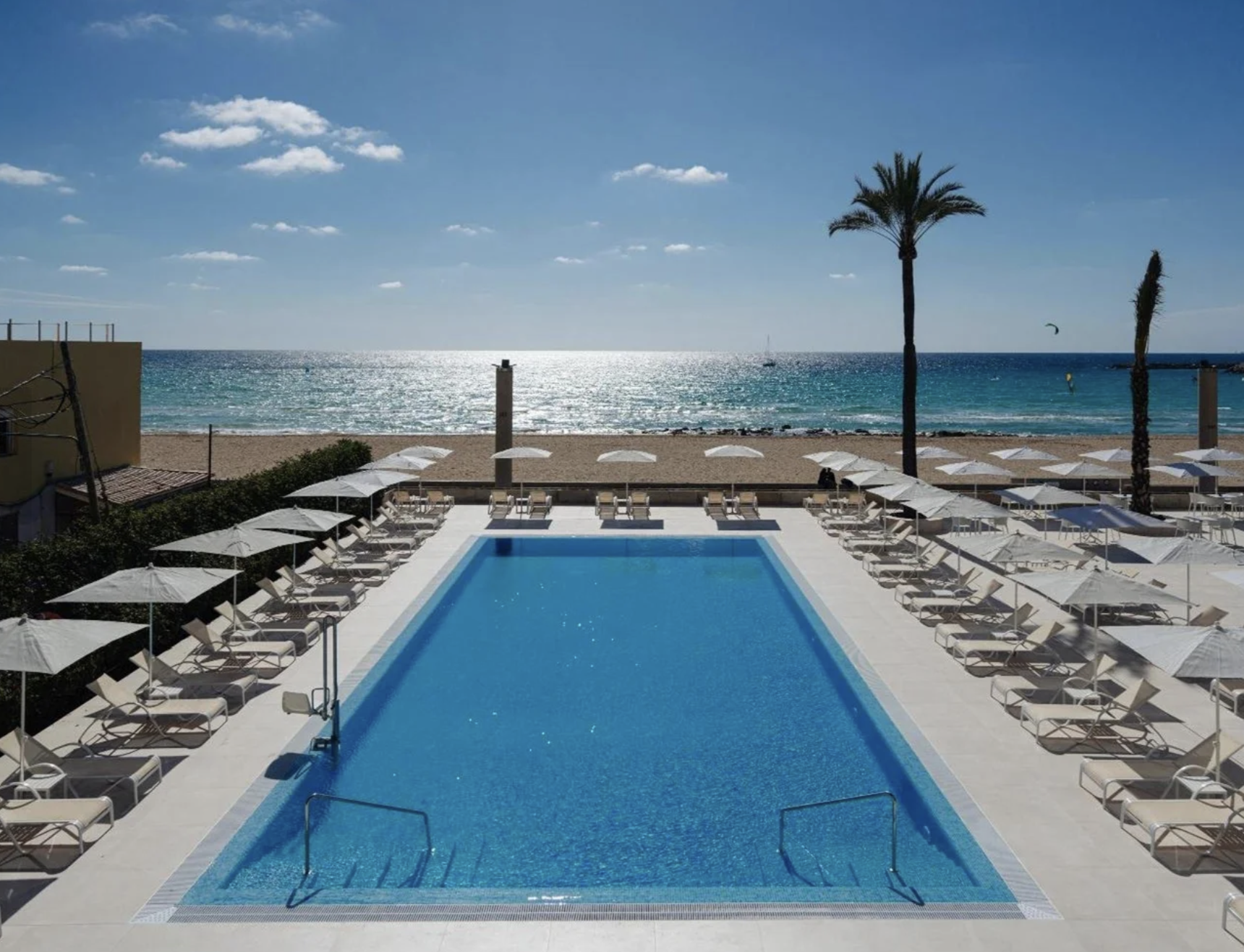 Piscina en un resort junto a la playa con sombrillas y sillas, vista al mar con palmeras y cielo despejado.
