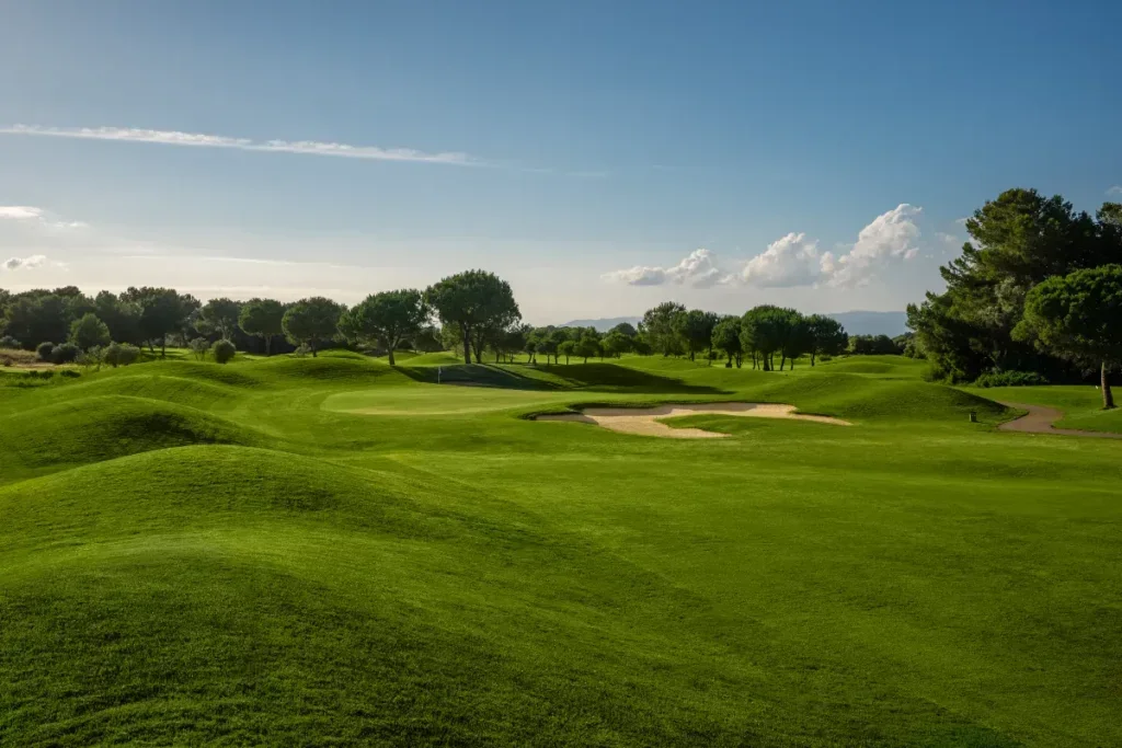 Campo de golf con césped bien cuidado, árboles y búnkeres de arena, bajo un cielo azul con nubes.