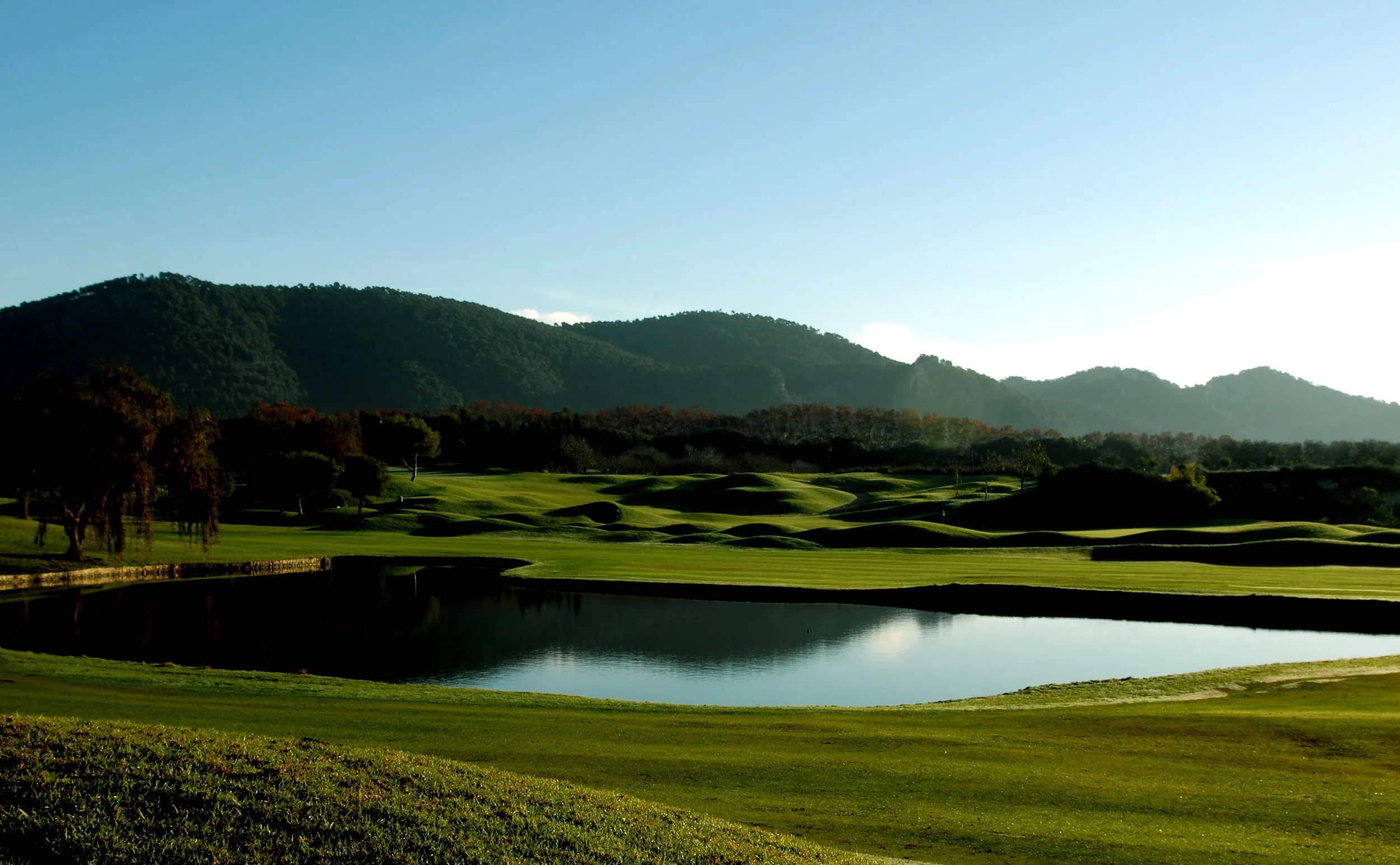 Campo de golf con césped verde, un lago y colinas en el fondo con árboles y montañas, bajo un cielo claro.