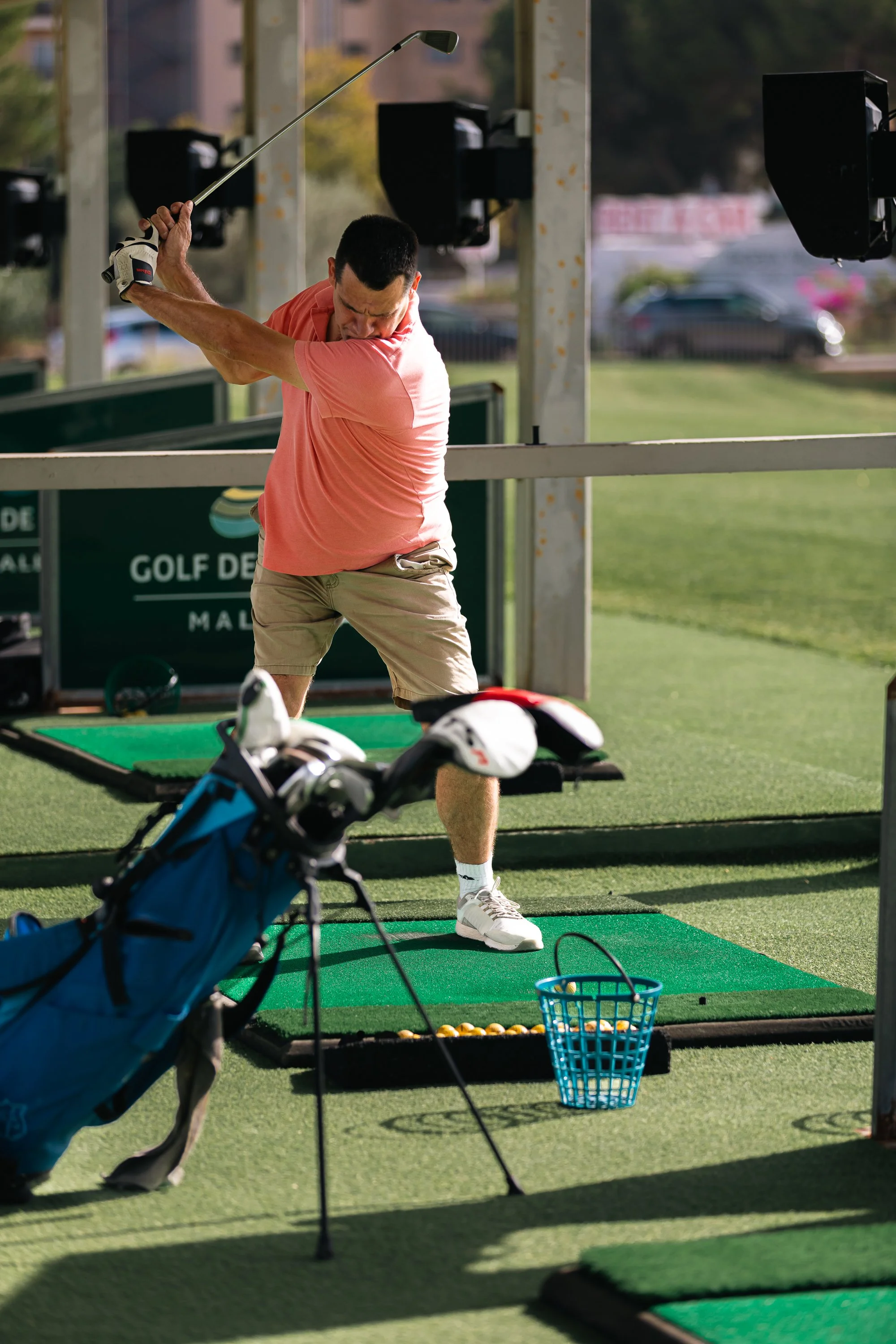Hombre practicando golf en un campo de prácticas, enmarcado con bolsas de golf y equipo de entrenamiento.