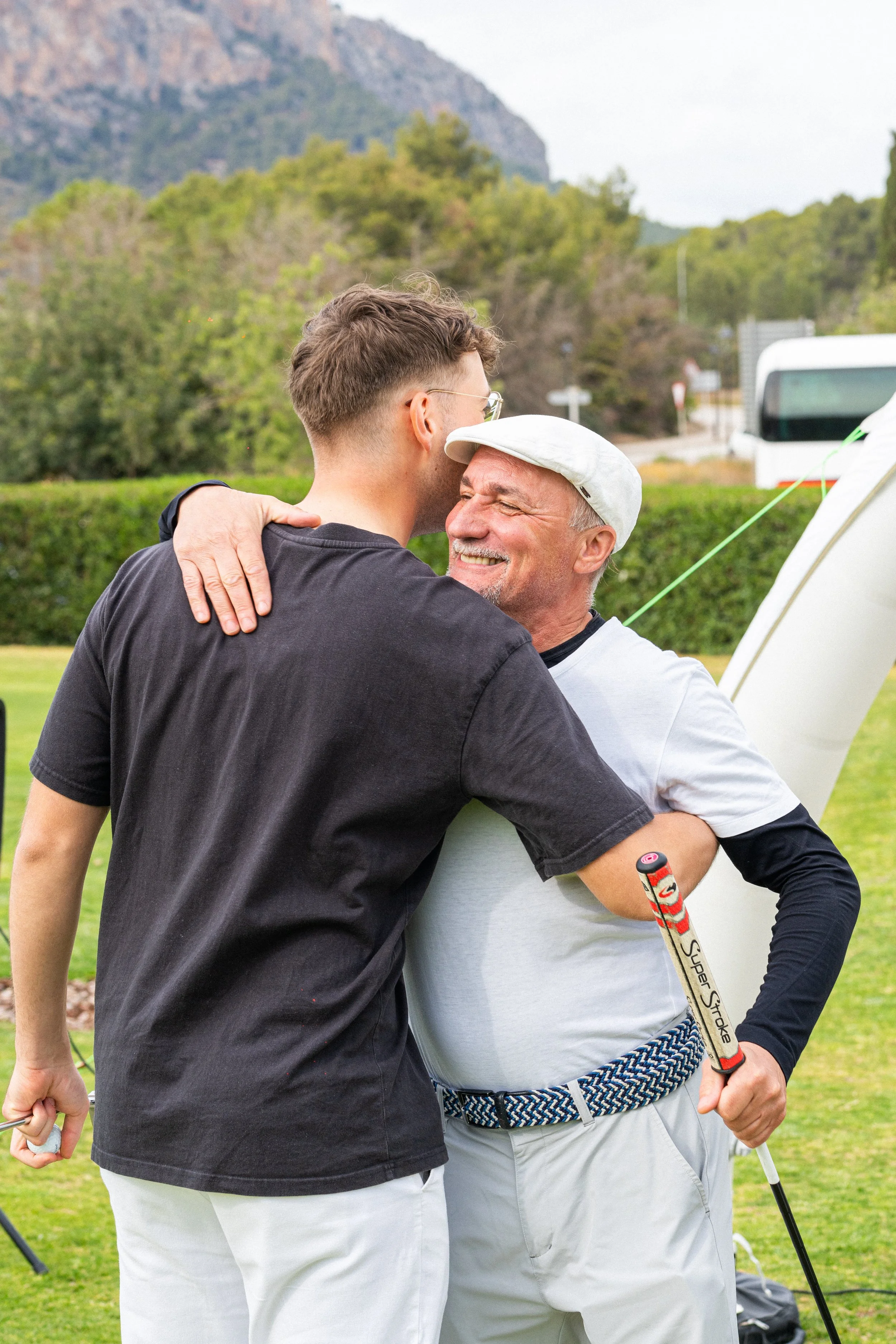 Un joven y un hombre mayor se abrazan en un campo de golf, ambos sonrientes.
