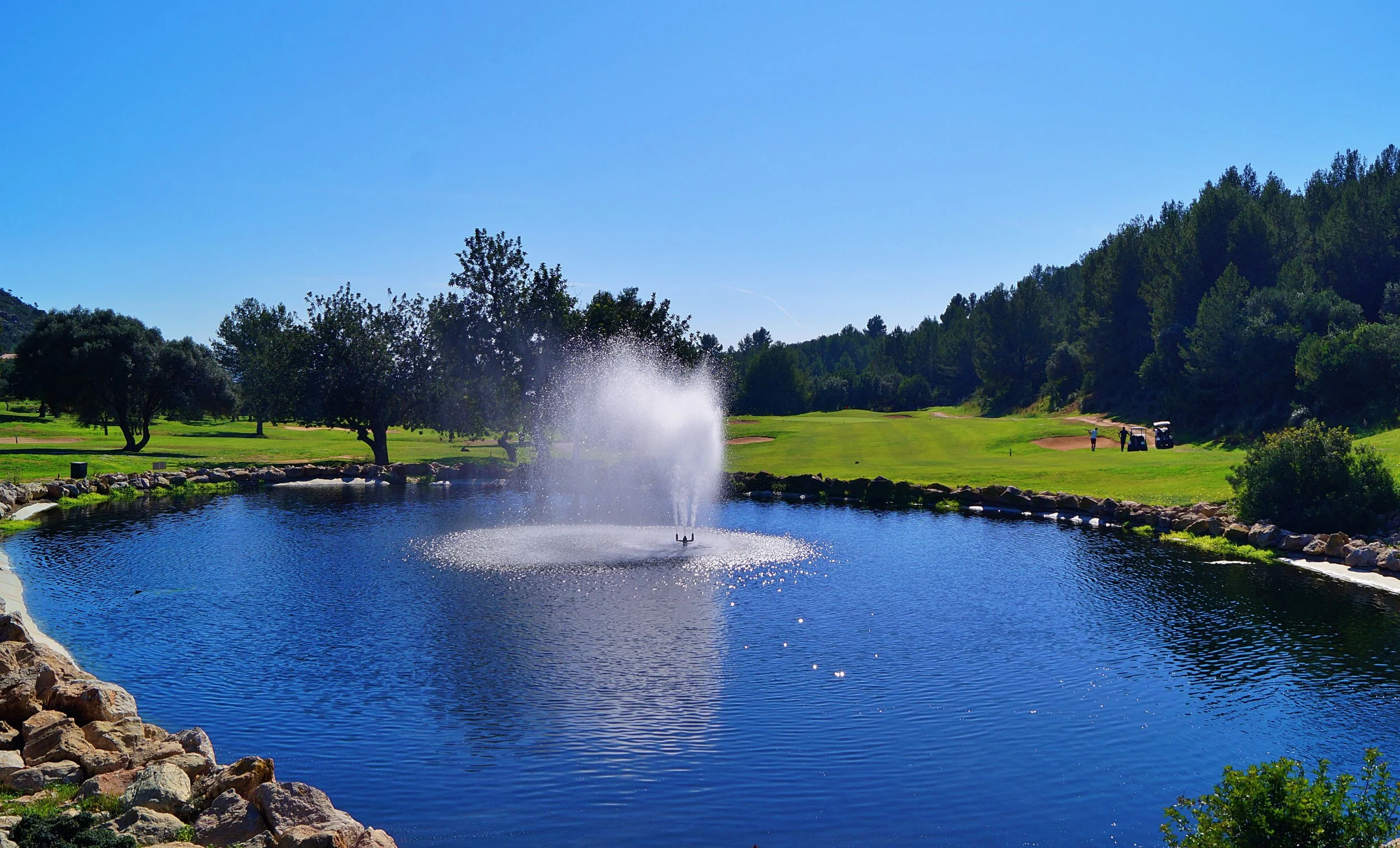 Campo de golf con lago y fuente, árboles y toques de cabaña en el fondo, día soleado con cielo despejado.