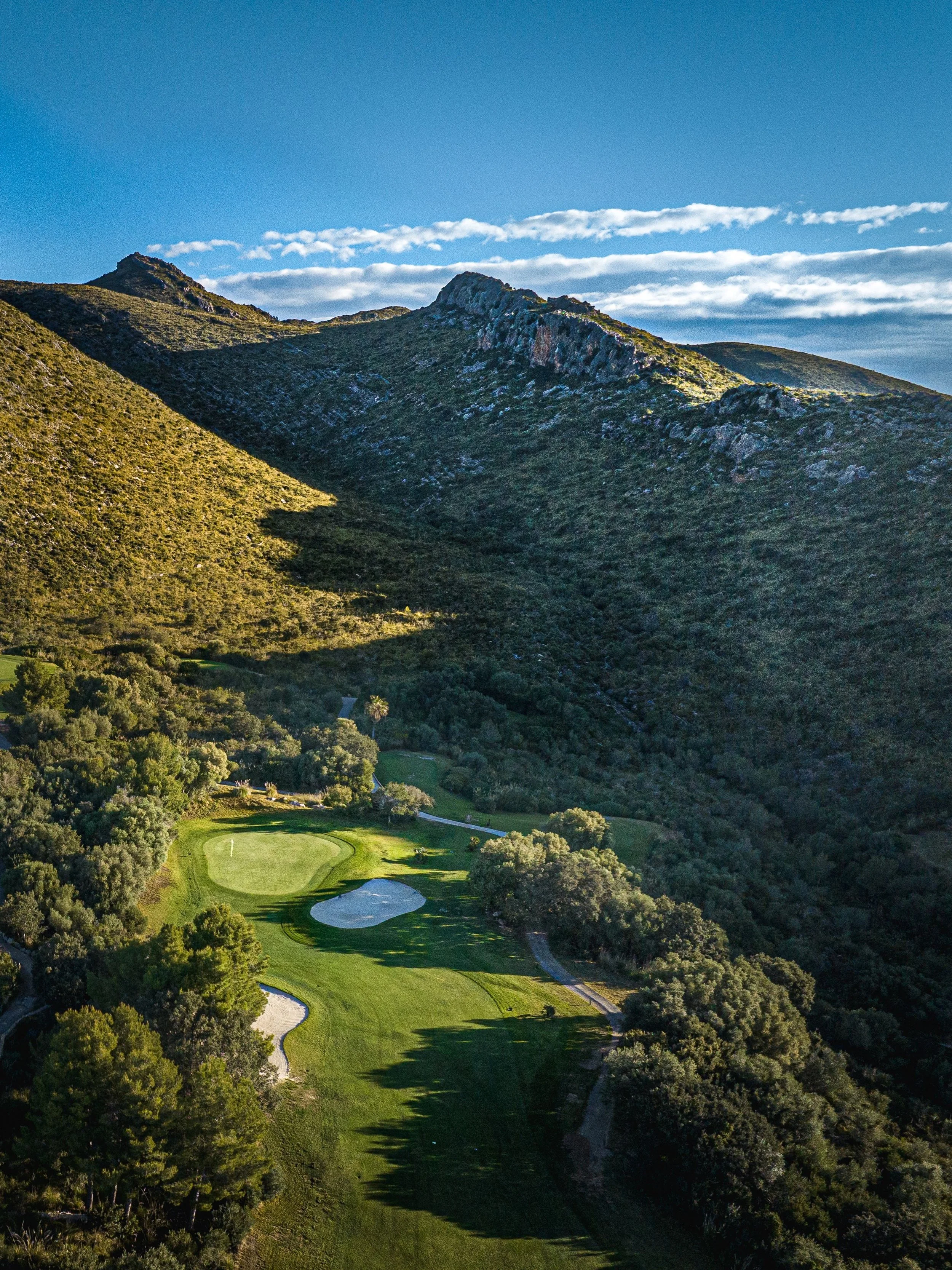 Paisaje de montañas con vegetación y un campo de golf en primer plano, con un cielo despejado y algunas nubes delgadas.